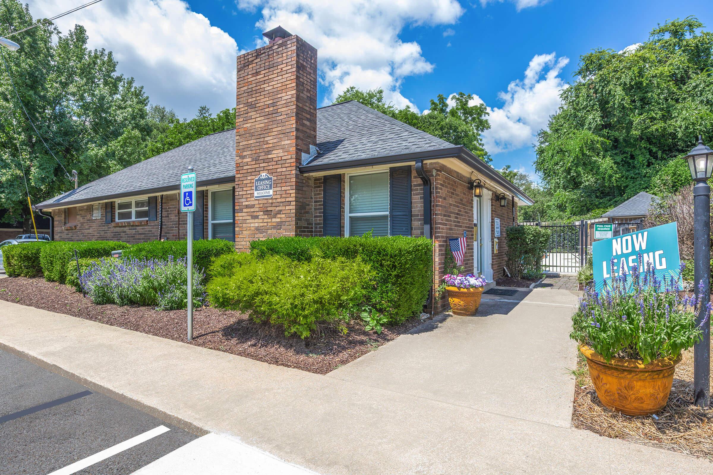 A single-story brick building surrounded by greenery, featuring a chimney. There are flower pots by the entrance and a blue sign that reads "NOW LEASING." The driveway is paved, and the scene is set against a backdrop of trees and a partly cloudy sky.