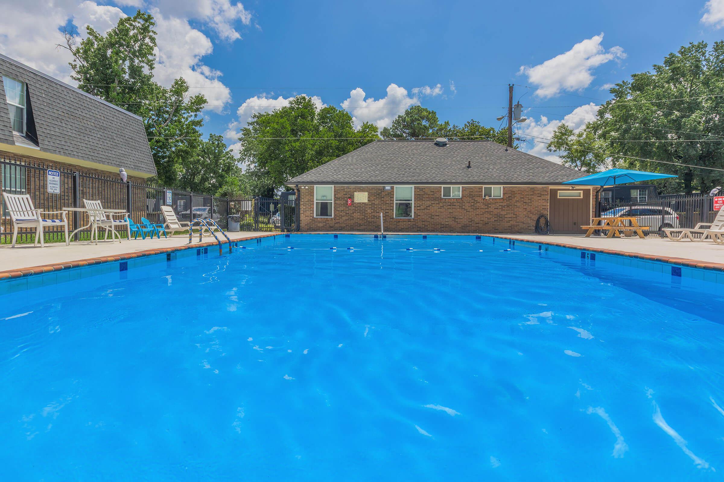 A clear blue swimming pool with lounge chairs along the side, surrounded by greenery and a nearby brick building. The sky is bright with scattered clouds, creating a sunny, inviting atmosphere. An umbrella is set up for shade, enhancing the relaxation space.
