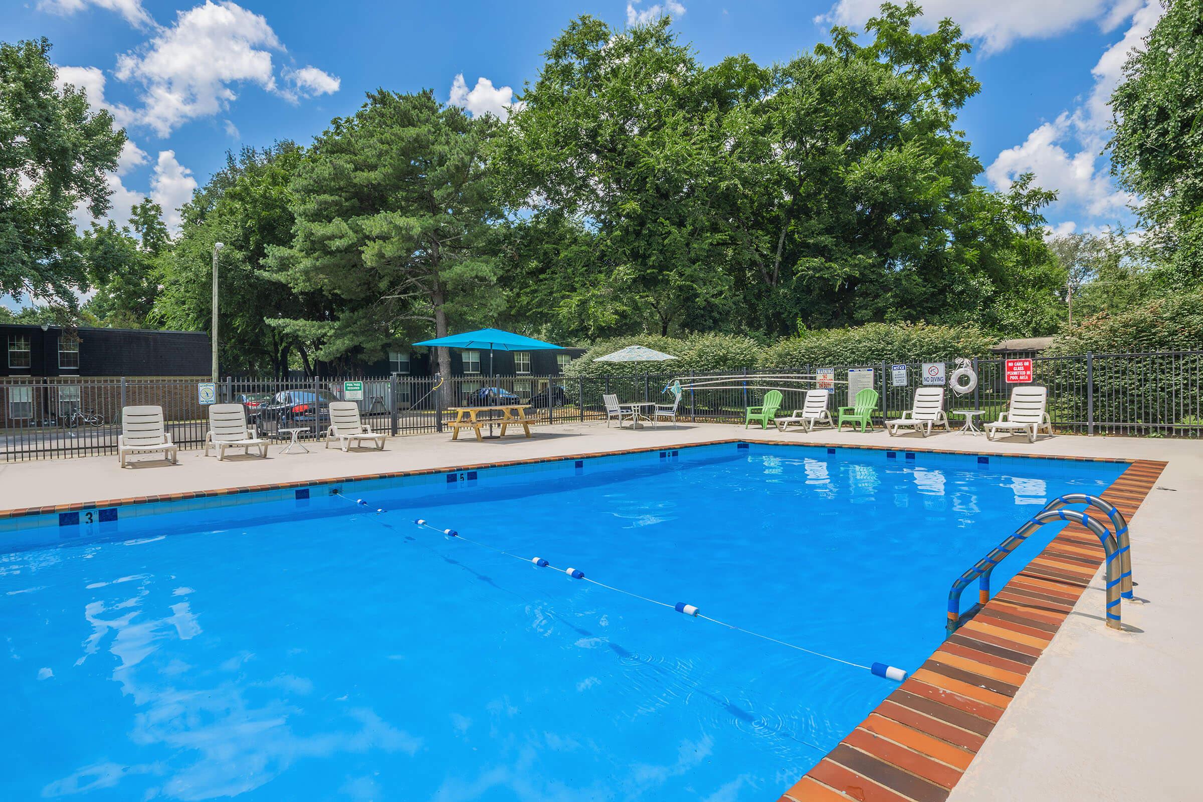 A bright, inviting swimming pool surrounded by lounge chairs and picnic tables, set in a lush green environment with trees and blue sky above. The pool features clear blue water and safety signage along the fence, creating a relaxing outdoor space for leisure and enjoyment.