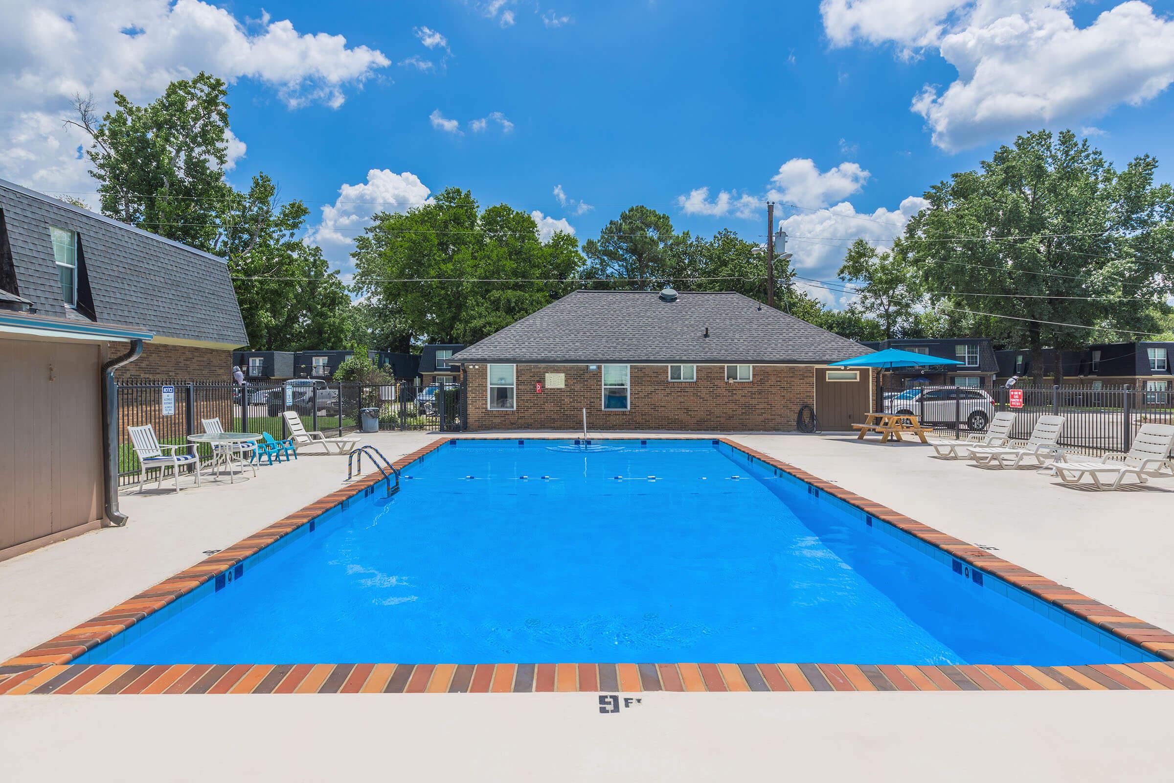 A bright, outdoor swimming pool surrounded by a concrete deck and lounge chairs. The pool is filled with clear blue water, and a few trees and buildings are visible in the background under a partly cloudy sky. Sunlight reflects off the water, creating a welcoming atmosphere for relaxation and leisure.