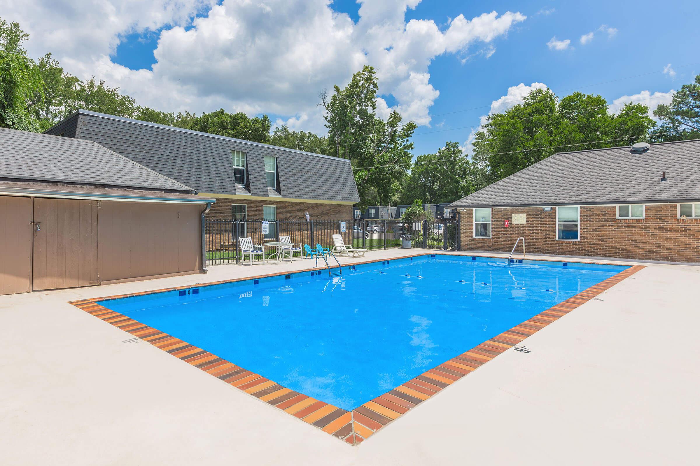 A vibrant outdoor swimming pool surrounded by a concrete deck, with lounge chairs scattered around. Nearby, two brick apartment buildings with trees in the background under a partly cloudy blue sky. The pool features a diving board and a ladder for access.