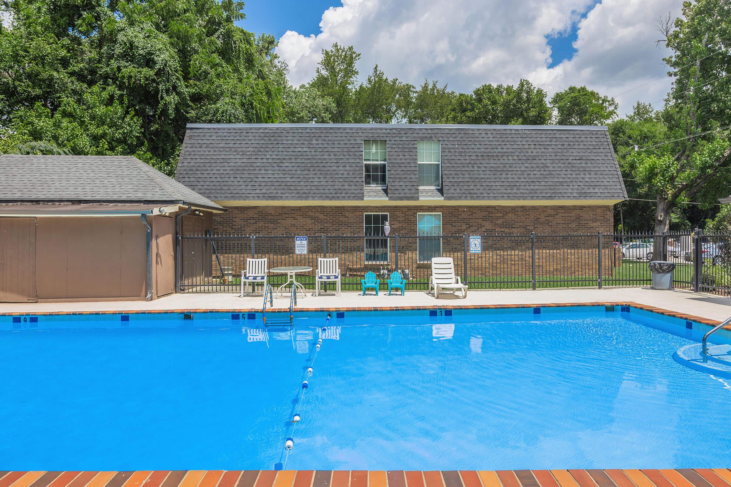 A swimming pool with clear blue water is surrounded by a fence. In the background, there is a brick building with several windows and a flat roof. Poolside, there are white lounge chairs and two blue chairs. The scene is under a bright sky with some clouds and greenery in the background.