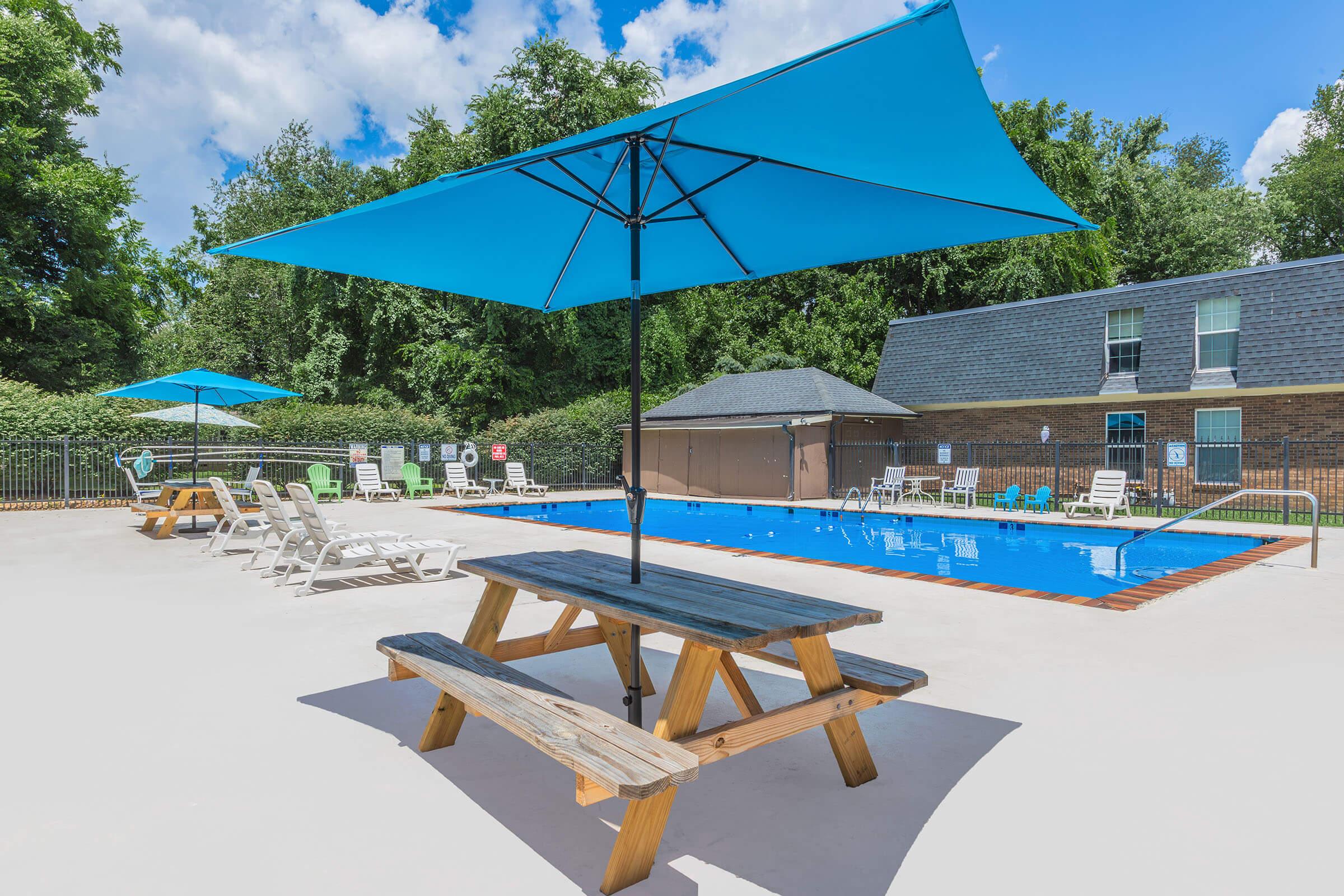 A spacious outdoor swimming pool area featuring a clear blue pool surrounded by lounge chairs and umbrellas. A wooden picnic table sits in the foreground, providing a shaded spot for relaxation. Lush greenery and a building are visible in the background under a bright blue sky with fluffy clouds.