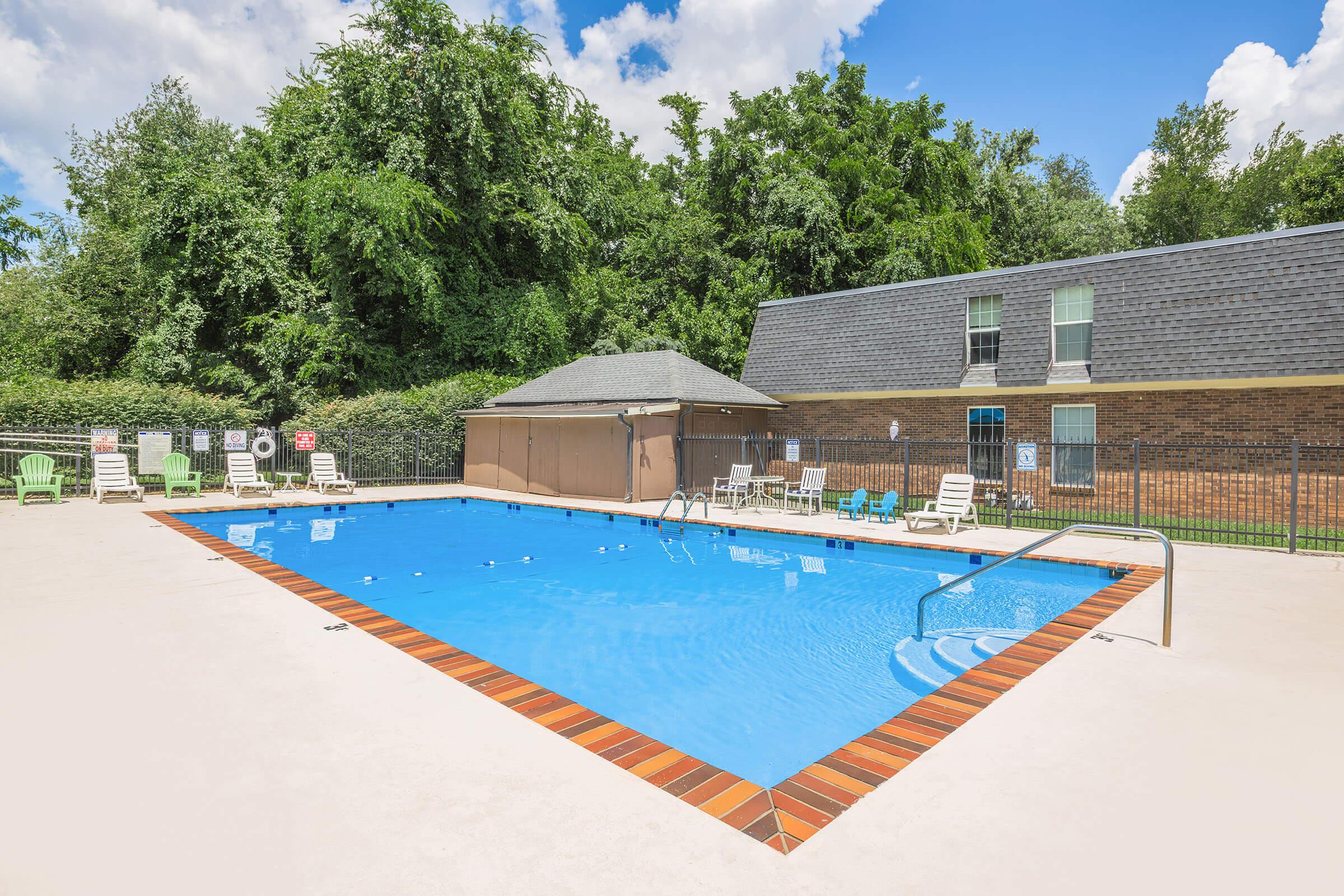 A bright blue swimming pool surrounded by lounge chairs, with a backdrop of lush green trees. The pool area features a fenced perimeter, and a small building can be seen nearby. The sky is clear with a few white clouds, creating a pleasant atmosphere for relaxation.