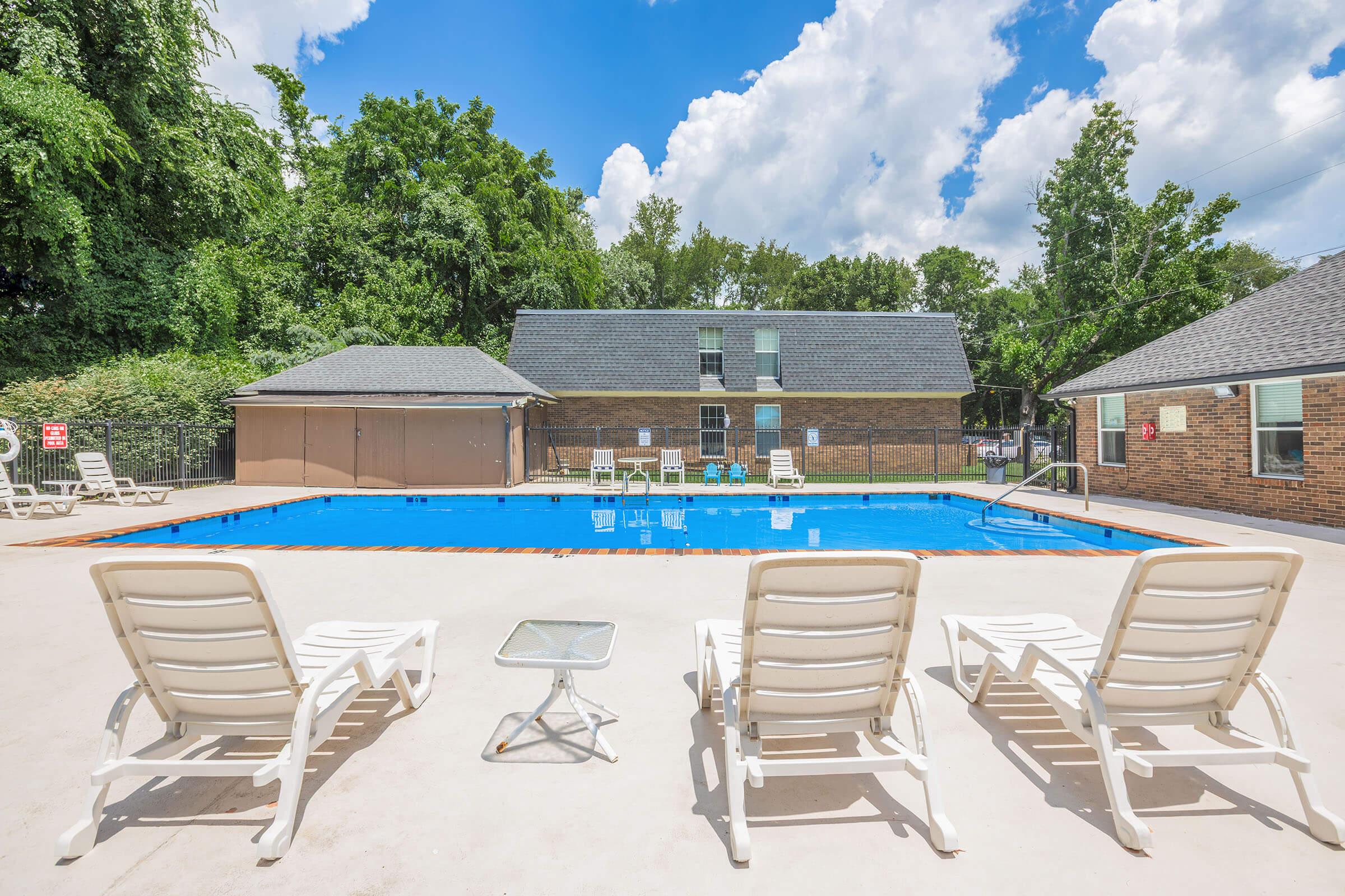 A bright outdoor pool area featuring a clean blue swimming pool surrounded by lounge chairs. In the background, there are two buildings with dark roofs and greenery lining the perimeter. The sky is clear with a few fluffy clouds, creating a pleasant atmosphere for relaxing by the pool.