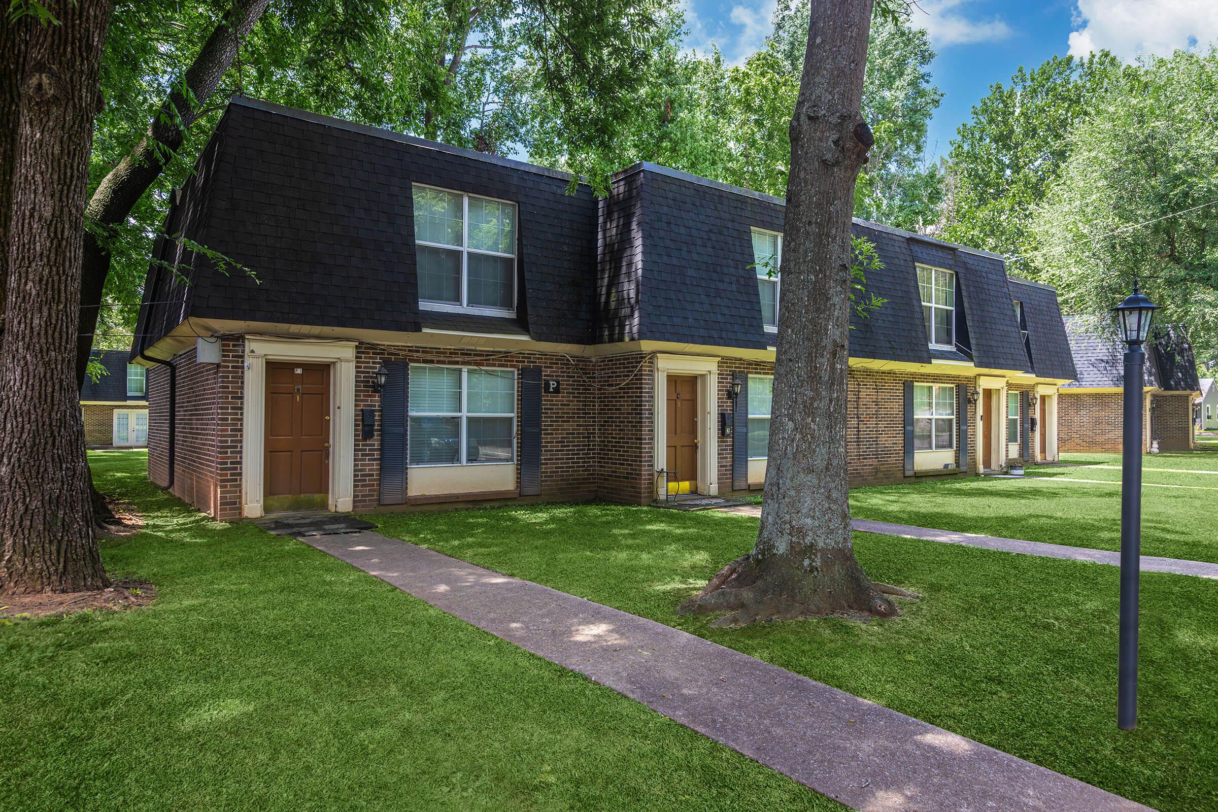 Two-story brick apartment buildings with dark shingled roofs, surrounded by green lawns and trees. A light post stands near a walkway leading to the entrance of each unit. The sky is blue with a few clouds, creating a bright and inviting atmosphere.