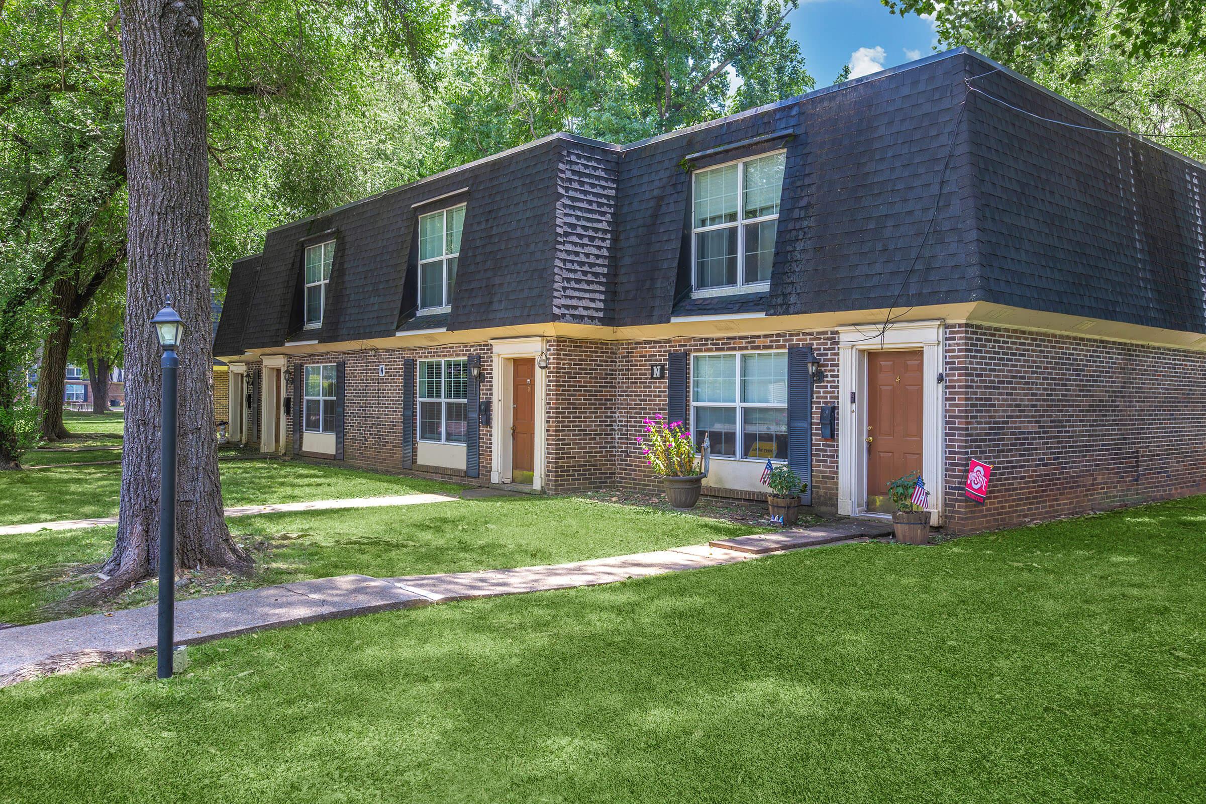 A multi-unit residential building with a brick exterior and dark shingles, set in a green landscape. The building features several doors, flower pots by the entrances, and is surrounded by trees. A lamp post stands nearby on a walkway leading to the building.