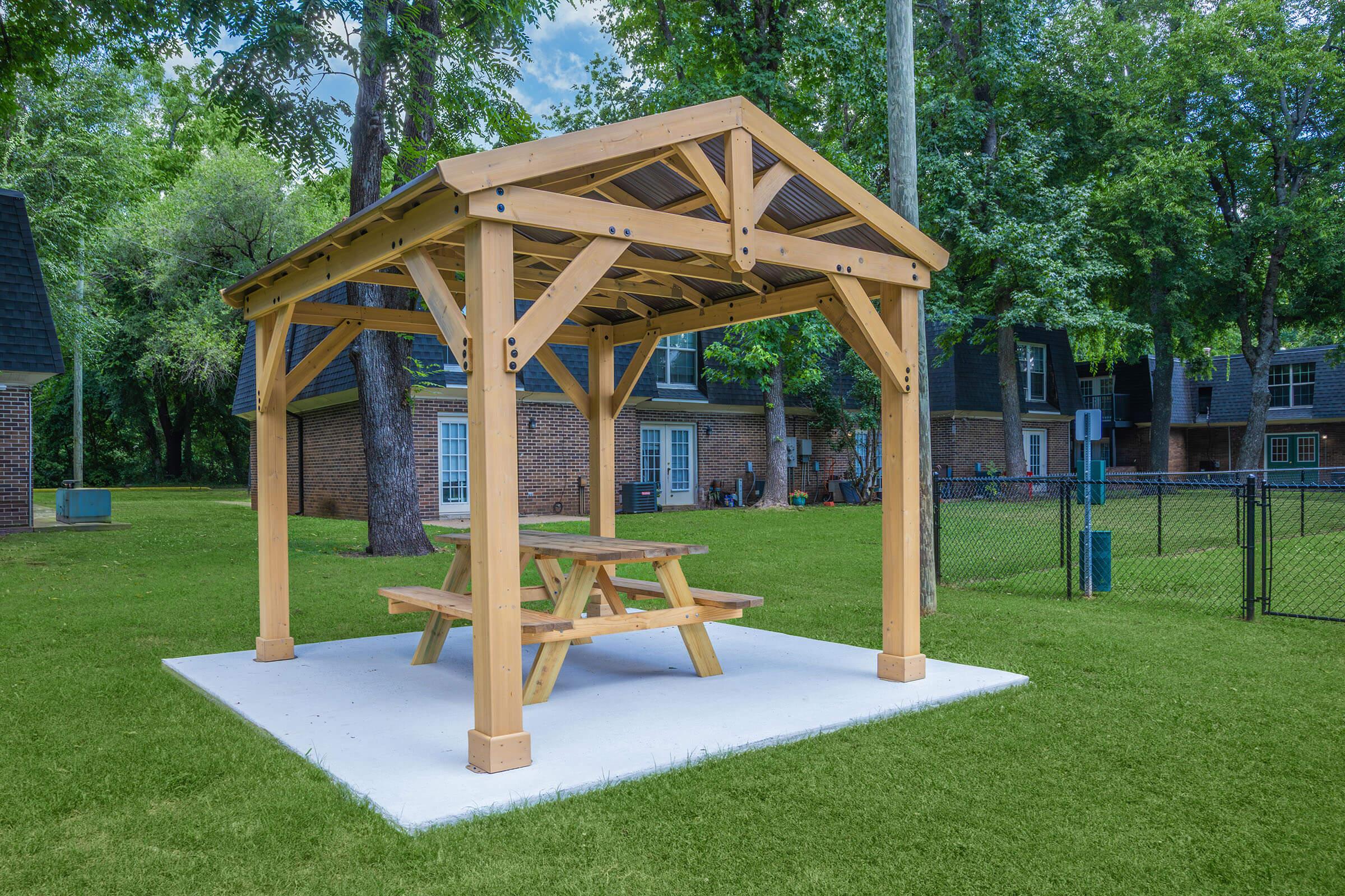 A wooden gazebo with a picnic table underneath, situated on a concrete slab. The gazebo features a pitched roof and is surrounded by green grass and trees, with buildings visible in the background. The scene suggests an outdoor recreational area.