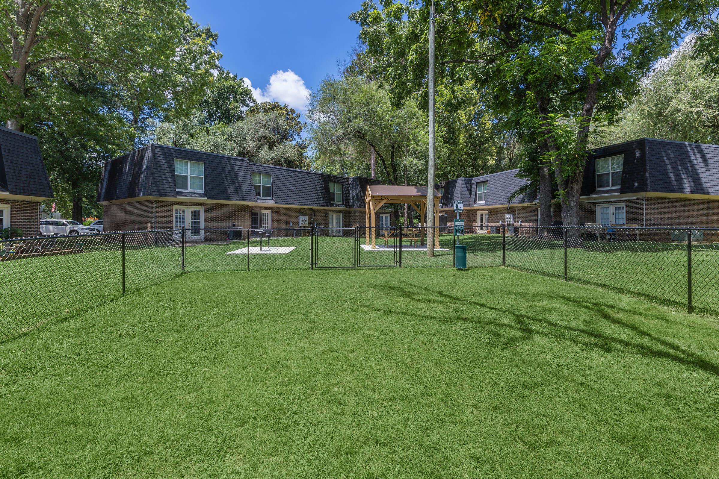 A landscaped area with green grass enclosed by a black fence, featuring two buildings with dark roofs. A small gazebo sits in the center, surrounded by trees under a clear blue sky, creating a peaceful setting for residents.