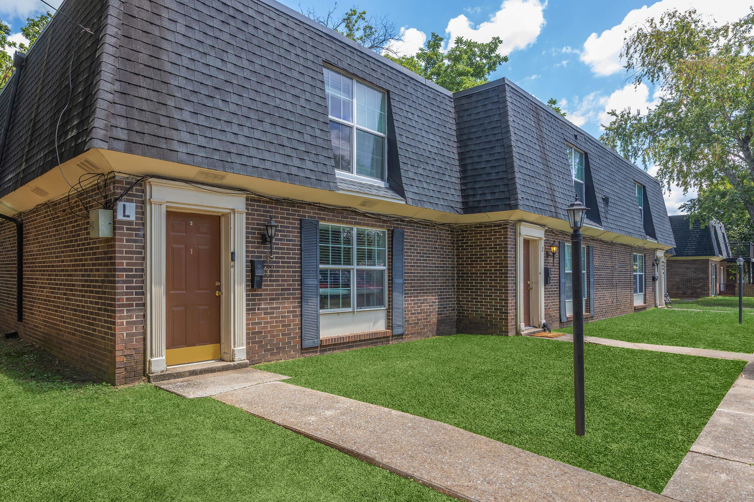 Two-story brick apartments with sloped roofs and large windows. Each unit has a front door with a small lamp post nearby. The pathway is lined with green grass, and trees are visible in the background under a partly cloudy blue sky.