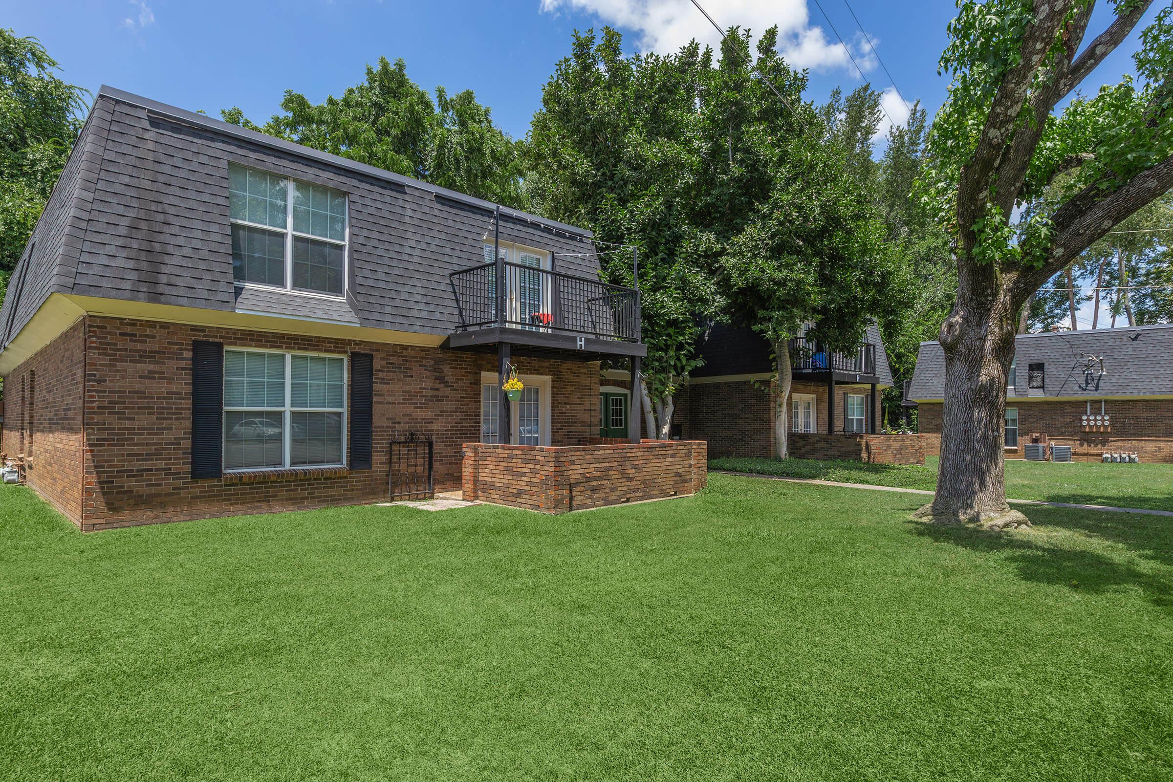 A view of a multi-unit brick building with a dark shingle roof, featuring balconies and surrounded by greenery. The lawn is well-maintained with lush grass, and there are trees providing shade in the area. The sky is clear with a few clouds, creating a bright and inviting atmosphere.