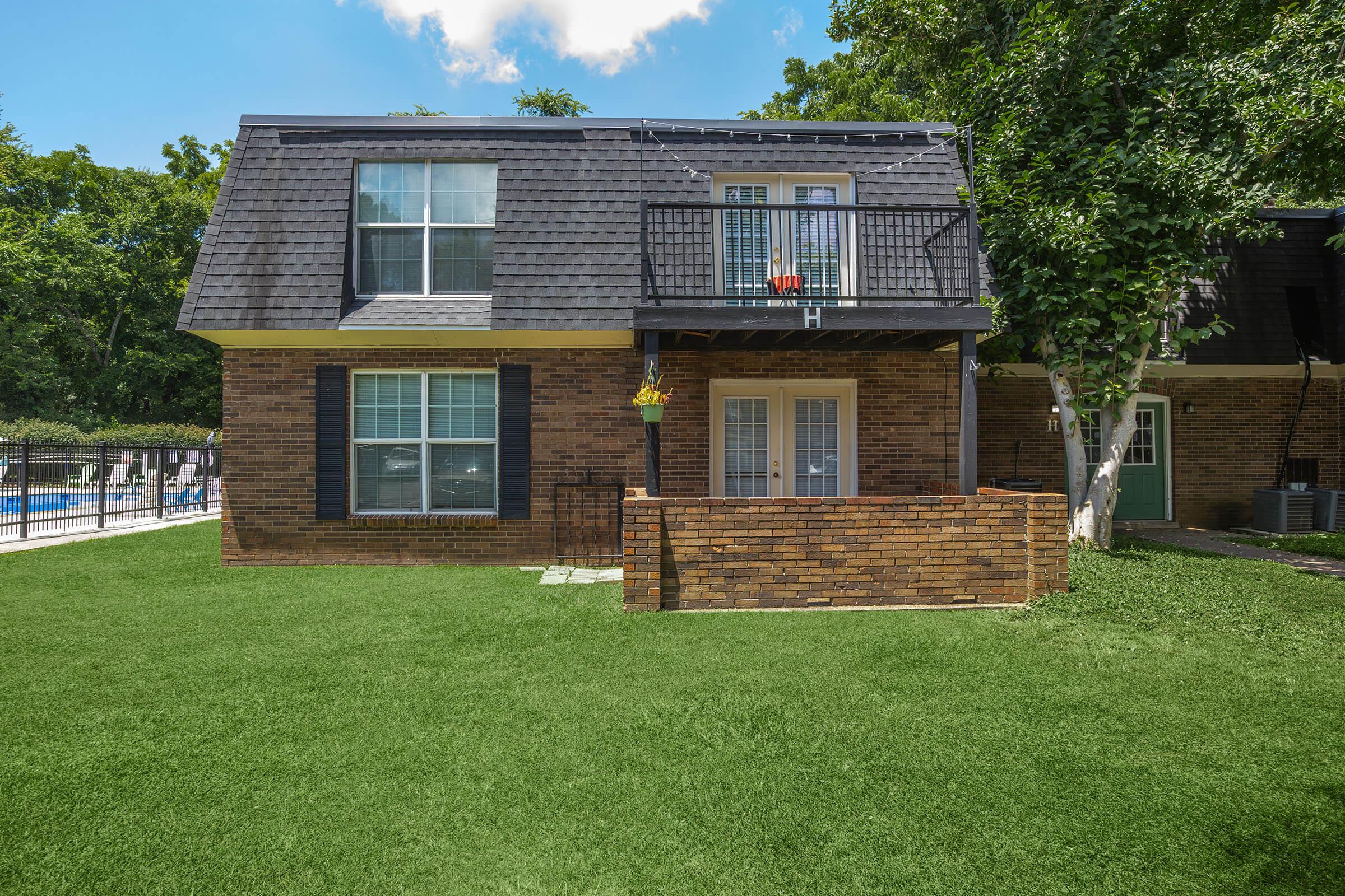 A two-story brick house with a gabled roof, featuring a balcony with a small hanging plant. The front yard is lush with green grass, and there's a fenced swimming pool visible in the background surrounded by trees. Bright blue sky with scattered clouds overhead.