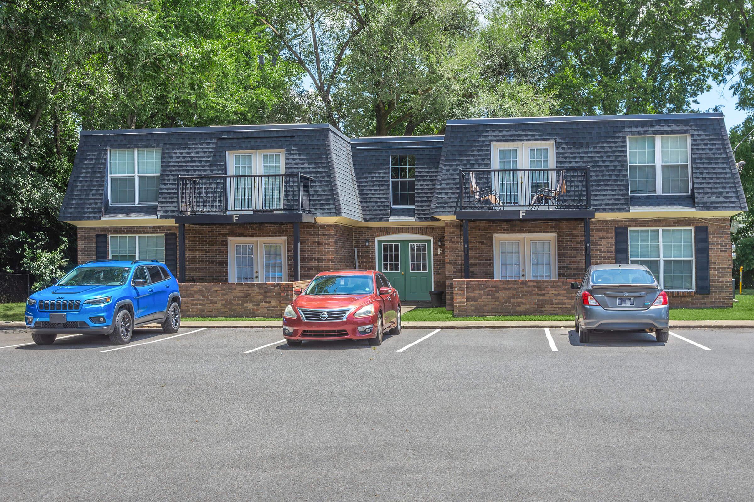 A two-story residential building featuring a dark shingle roof, balconies on the second floor, and a brick façade. Three cars—one blue SUV, one red sedan, and one silver sedan—are parked in front of the building on a sunlit day, surrounded by green trees in the background.