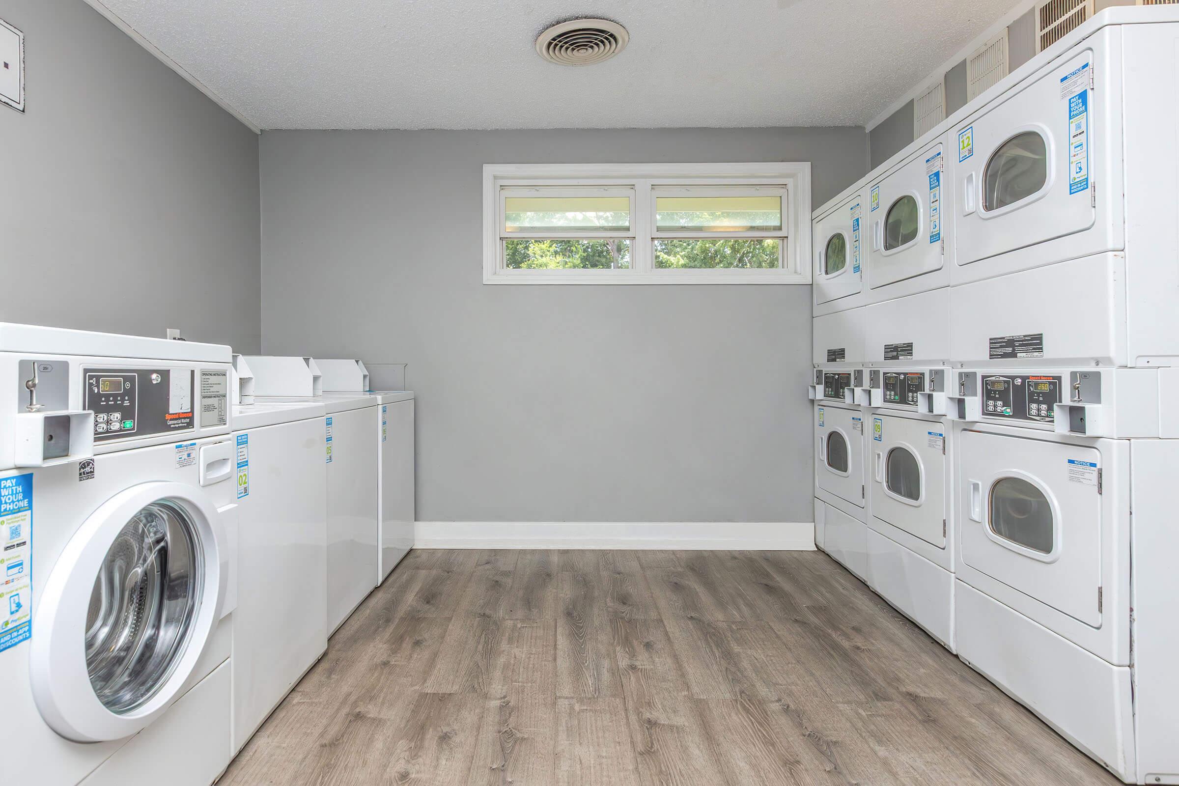 A clean, modern laundry room featuring several white washing machines and dryers lined up against light gray walls. Large windows allow natural light to fill the space, while the floor is covered with light wood-like laminate. The room is neat and organized, creating a functional environment for laundry tasks.