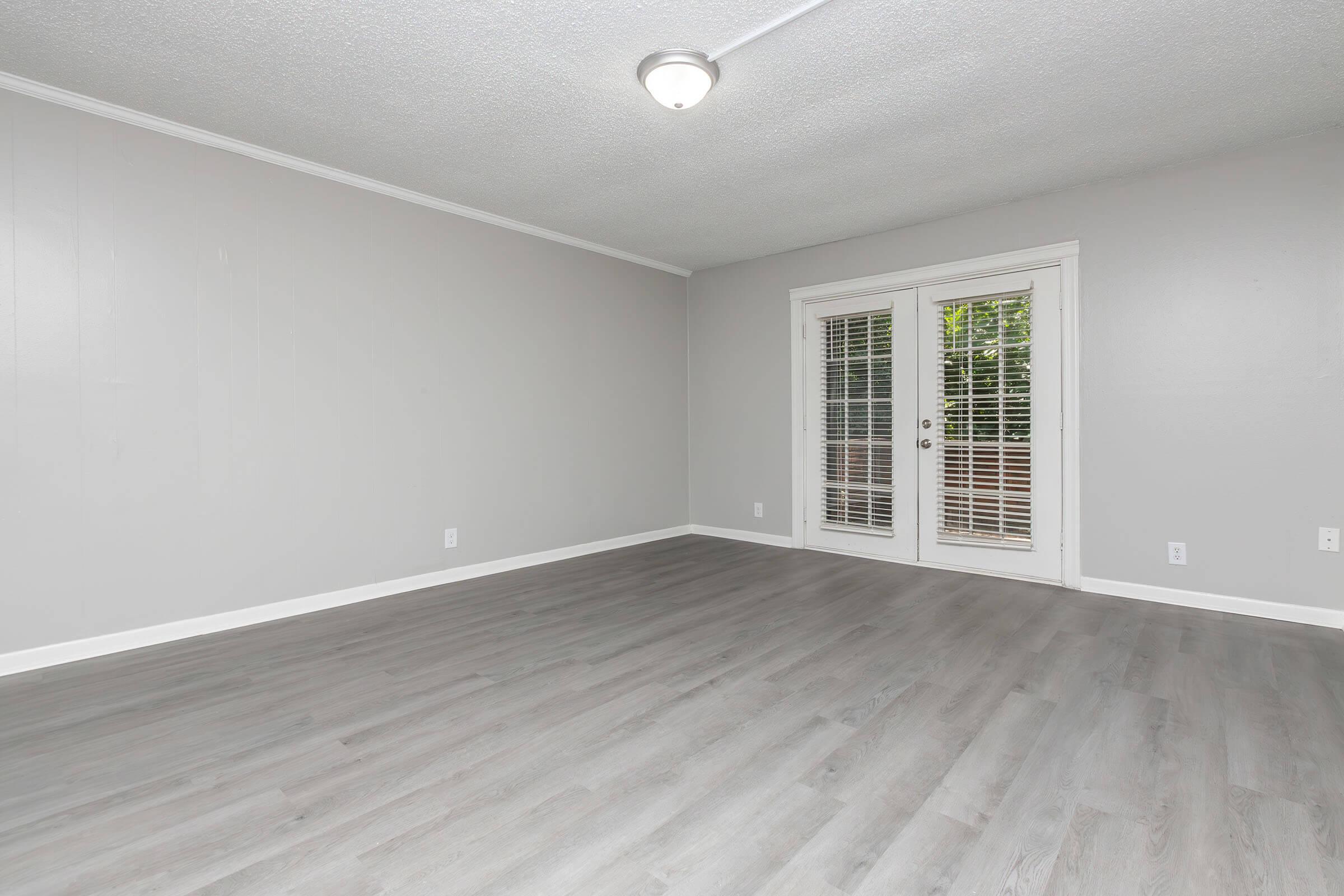 Empty room with light gray walls and flooring. A set of double doors with white blinds leads to an outdoor area. The ceiling features a simple light fixture, and the overall ambiance is bright and spacious, ideal for decorating.