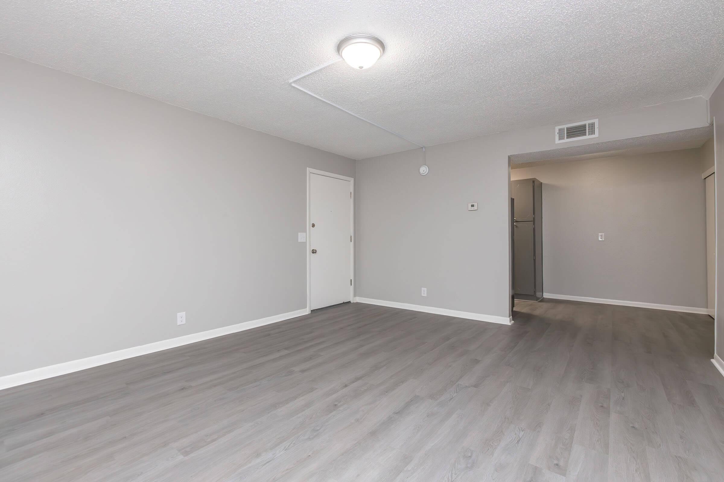 Empty room with gray walls and light wooden flooring. A ceiling light fixture is visible. There is a door on the left leading to another room, and a hallway leading to what appears to be a kitchen area in the background. The space is well-lit and appears freshly painted or renovated.