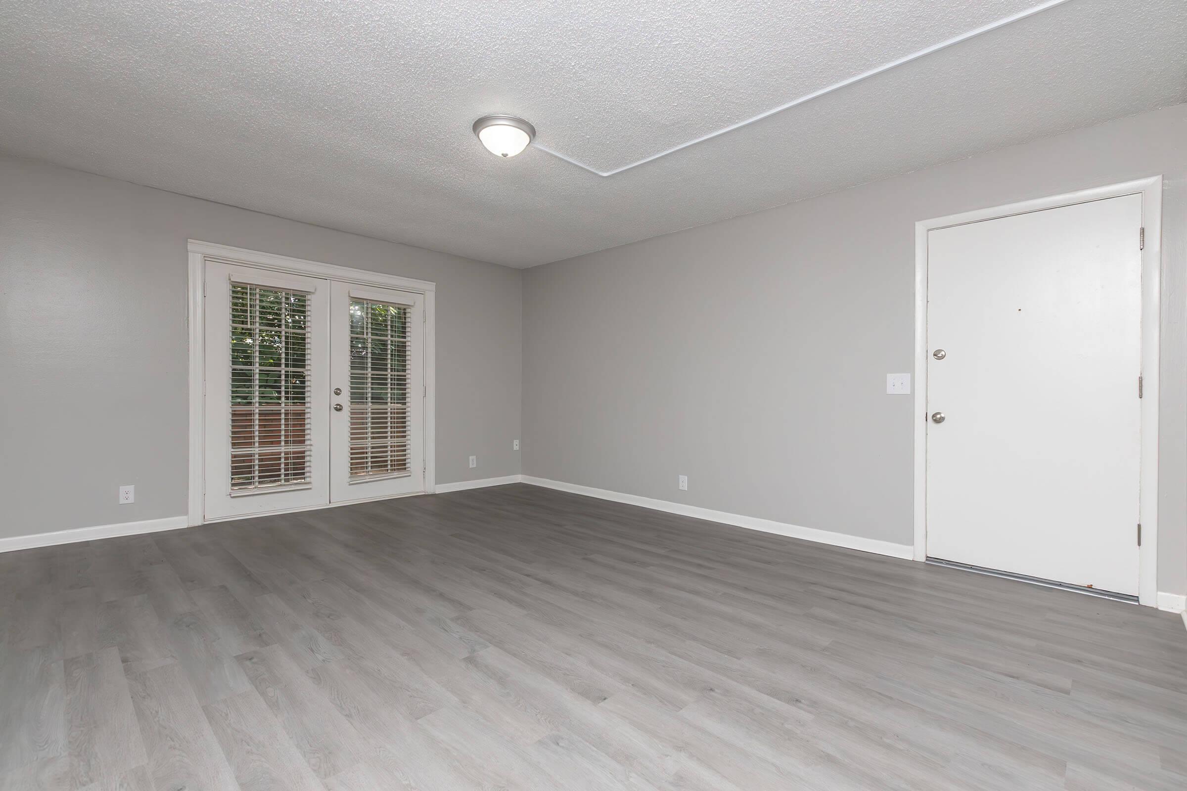 A spacious, well-lit room with light gray walls and a smooth, light-colored laminate floor. Two sets of French doors with blinds lead to an outdoor area, and a single door is present on one wall. The ceiling features a central light fixture, and the overall space appears empty and ready for furnishing.