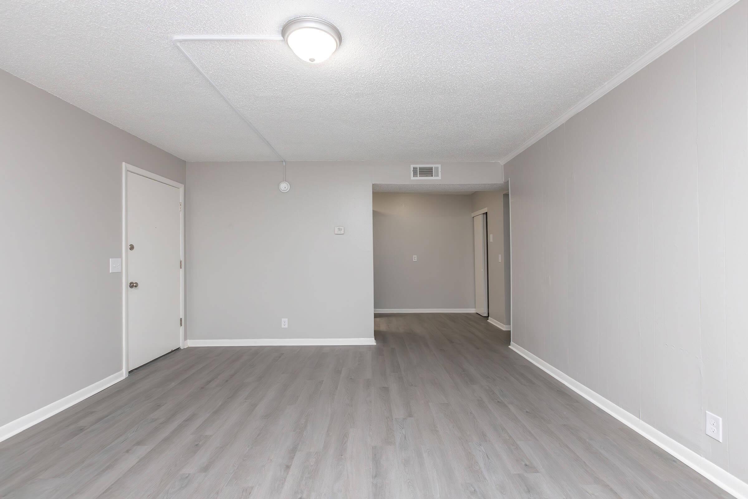 An empty room featuring light gray walls and a new laminate floor. A ceiling light fixture is visible, and there are doors leading to other spaces. The overall atmosphere is bright and spacious, with no furniture present.