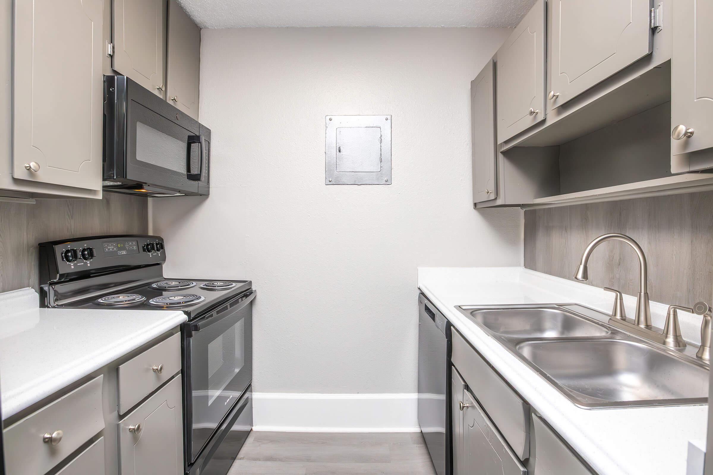 A modern kitchen featuring gray cabinetry, a microwave, an electric stove, and a double sink. The countertops are white, and the floor is light-colored. The walls are painted a soft gray, creating a bright and clean atmosphere. There is a small wall panel visible in the back.