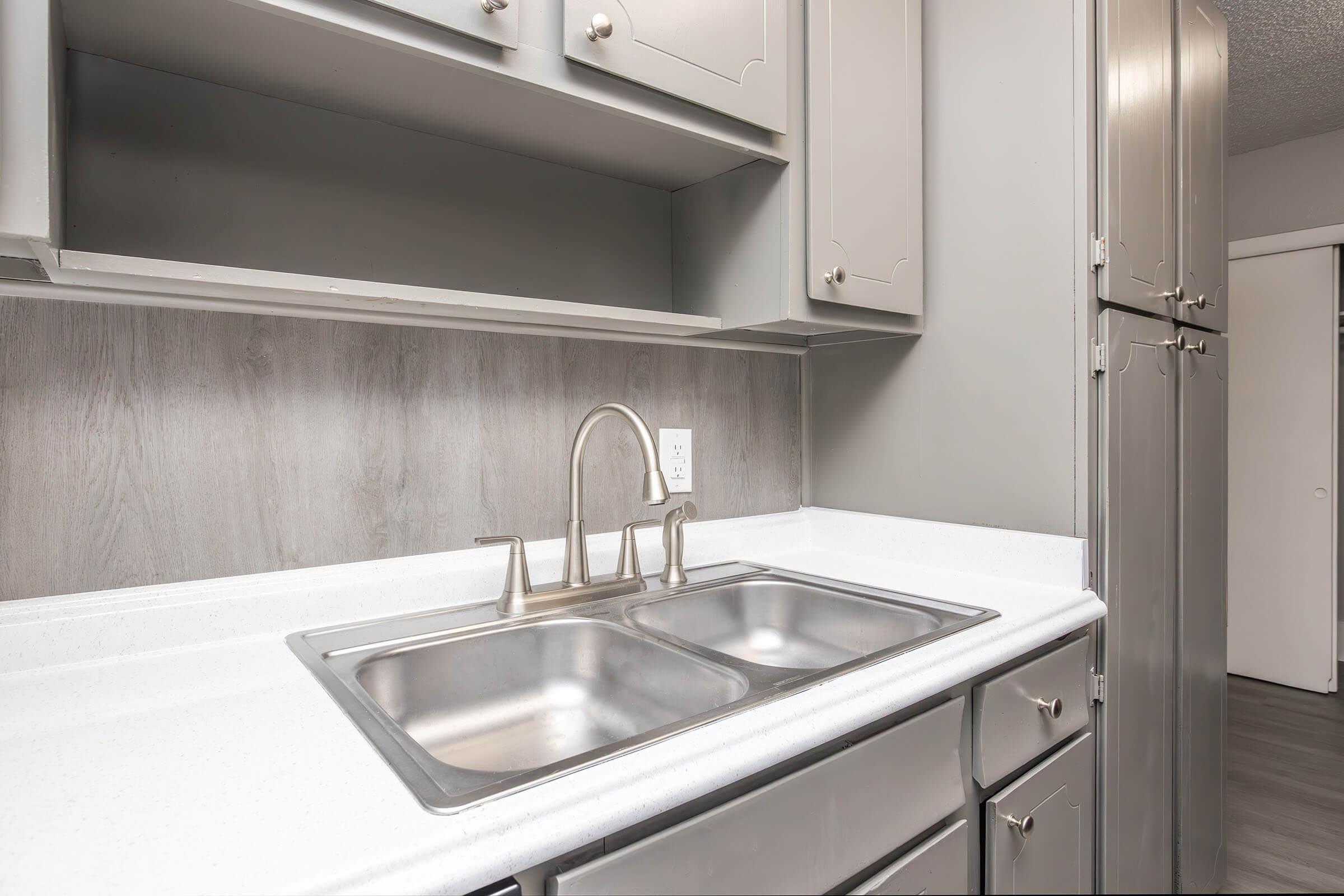 A modern kitchen featuring a white countertop with a double sink, gray cabinetry, and a stainless steel faucet. The walls are adorned with light gray paneling, and a refrigerator is visible in the background, along with a doorway leading to another room. The overall design is sleek and contemporary.