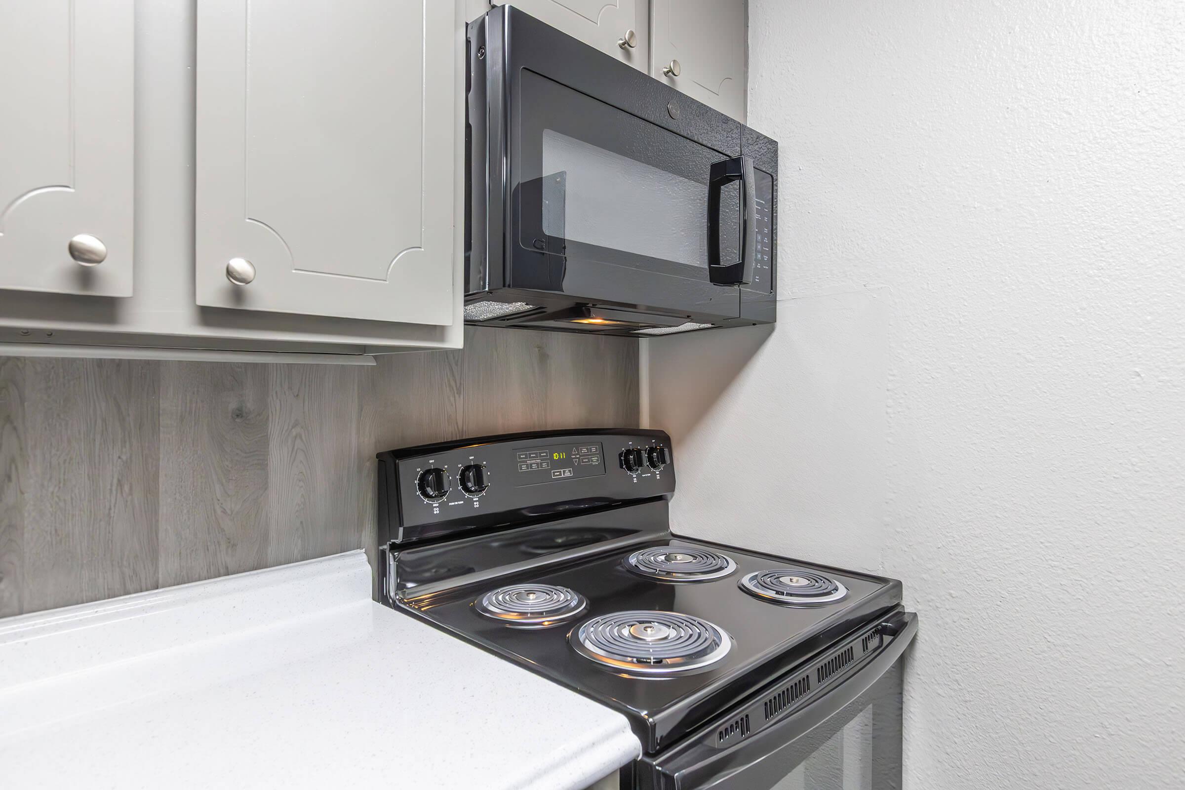 A modern kitchen setup featuring a black electric stove with four burners, a microwave mounted above, and light gray cabinets. The countertop is white, and the walls have a light wood paneling. The overall appearance is clean and contemporary.