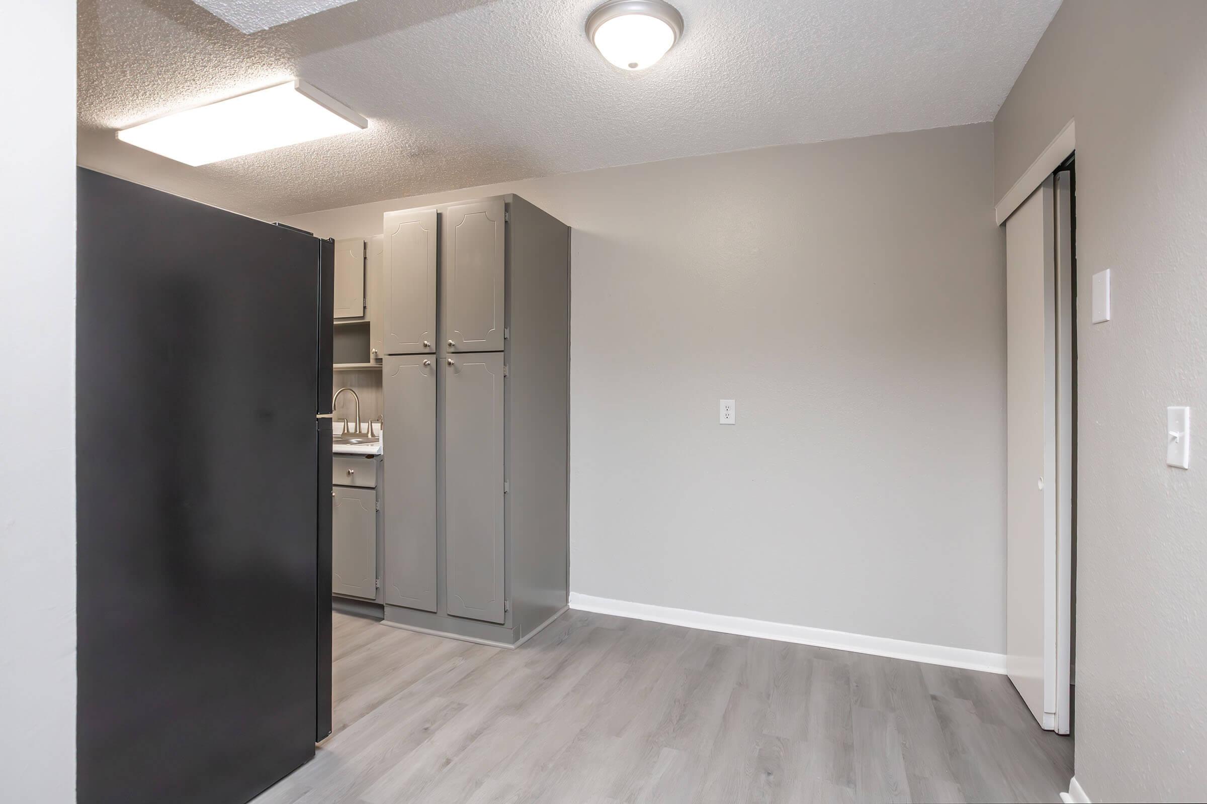 A small, modern kitchen space featuring a black refrigerator on the left, grey cabinets on the right, and a light fixture overhead. The walls are painted in a light grey, and the flooring appears to be a light wood laminate. The area is well-lit and organized, creating a clean and functional environment.