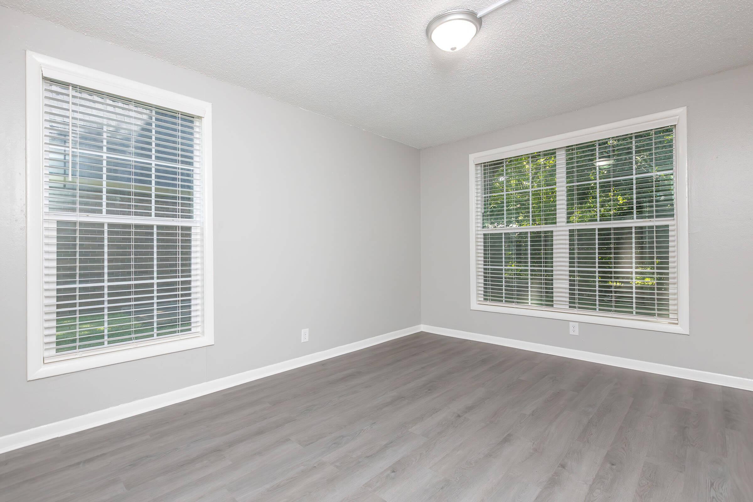 Empty room with light gray walls and hardwood floors. Two large windows with white blinds let in natural light. The ceiling light fixture provides additional illumination. The space has a minimalist feel, suitable for various uses.