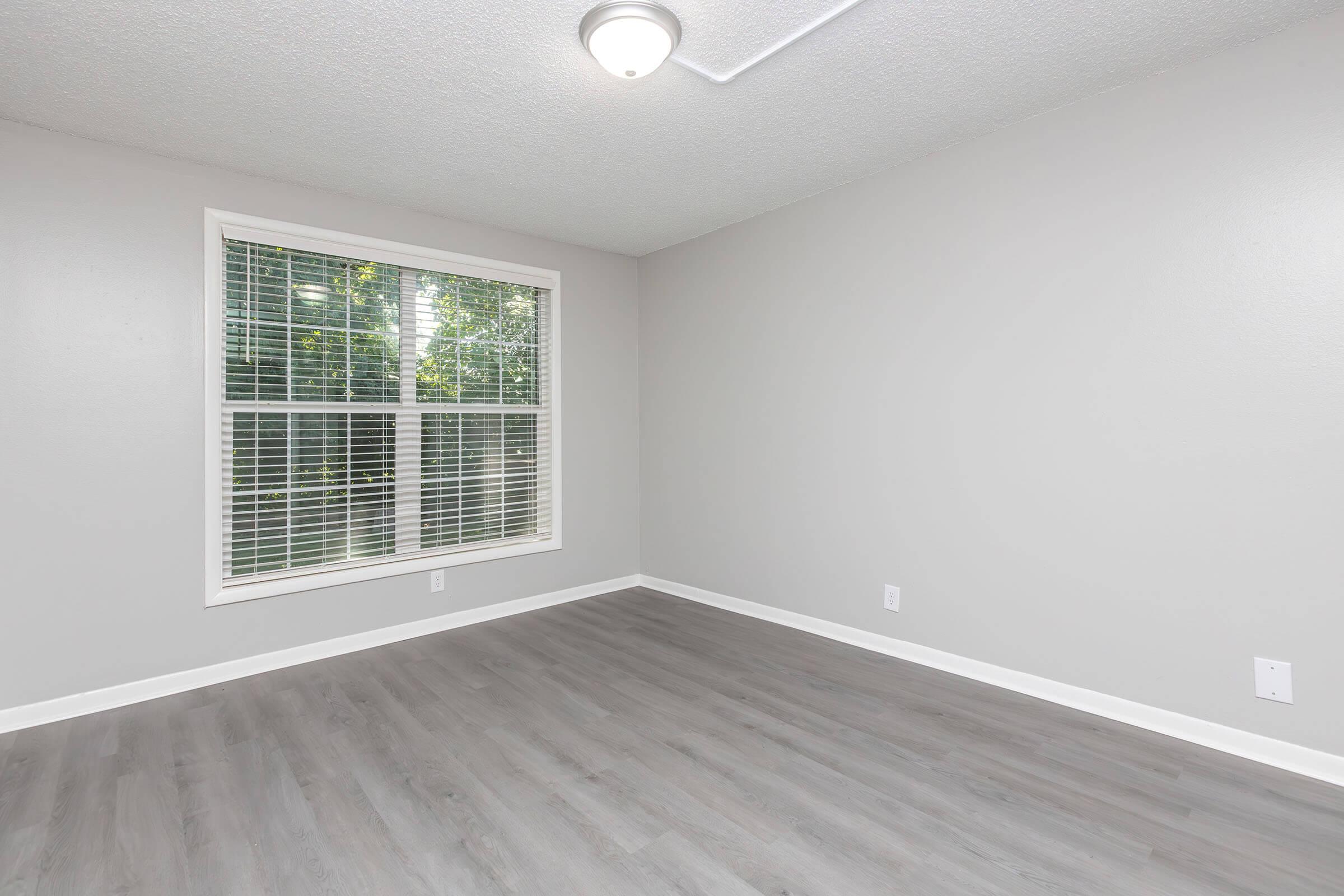Empty room with light gray walls and a smooth gray floor. A large window with white blinds allows natural light to enter, and there is a ceiling light fixture. The space appears clean and ready for furniture, with no decorations or personal items present.
