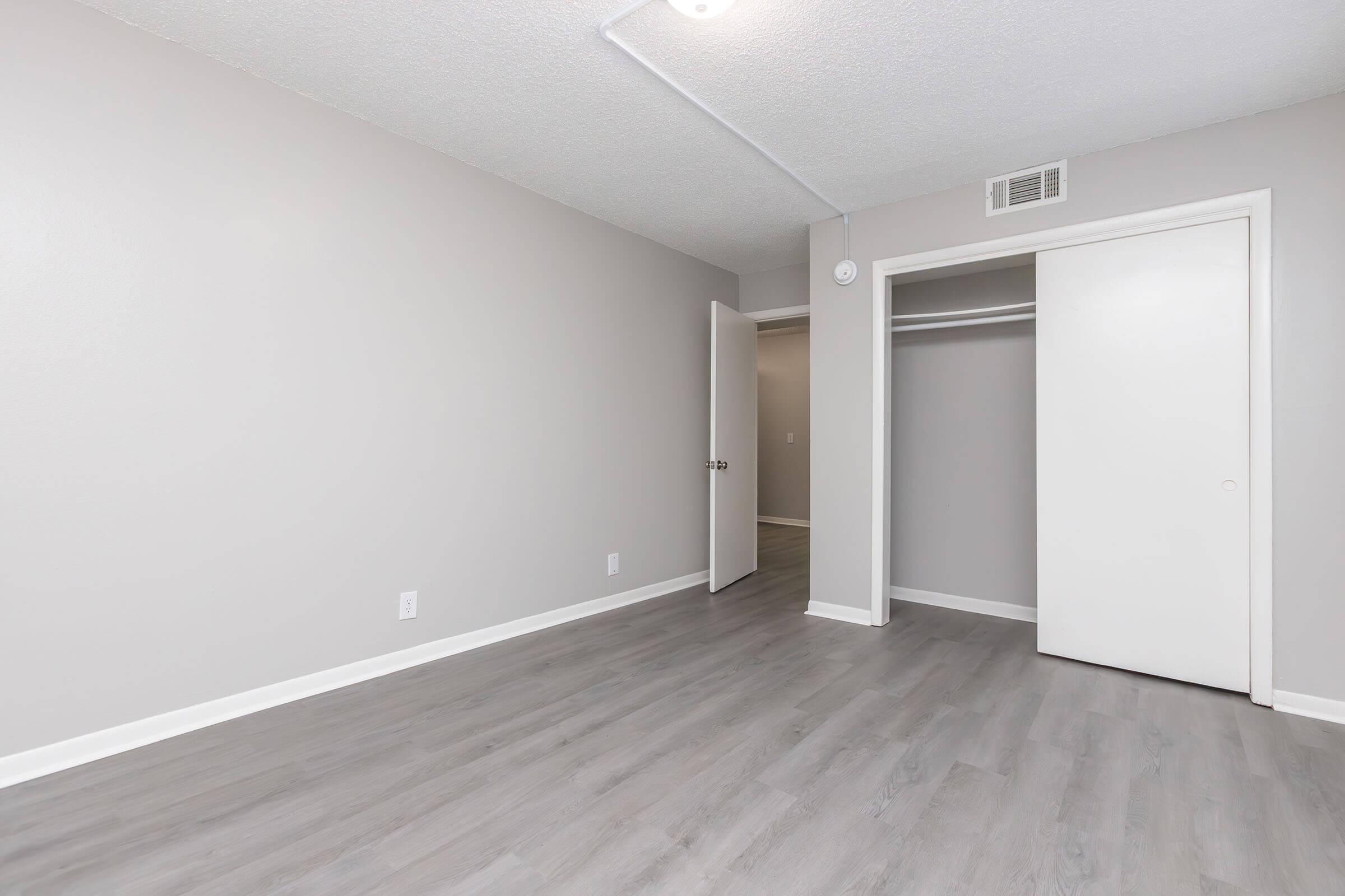 Empty room with light gray walls and a light gray laminate floor. A closed closet is visible with a sliding door, and an open door leads to another room. The ceiling is plain with a single overhead light fixture. The space appears clean and modern, suitable for various uses.