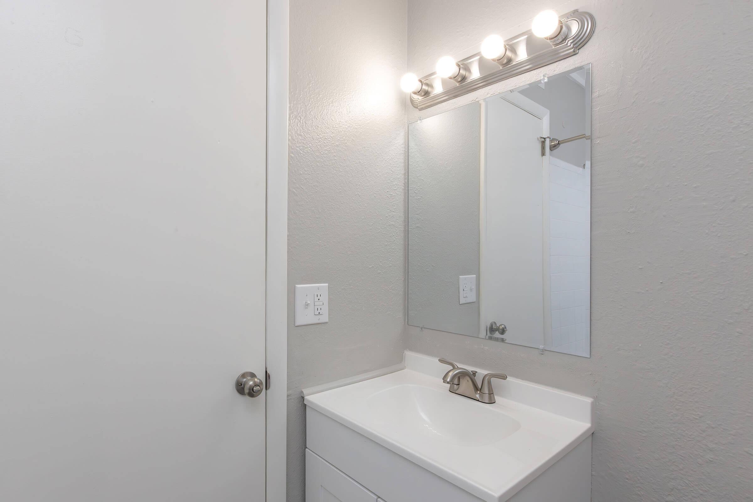 A modern bathroom featuring a white sink with a silver faucet, a large mirror above the sink, and a light fixture with four bulbs. The walls are painted gray, and there is a wooden door on one side. A shower can be seen in the background through an open door.
