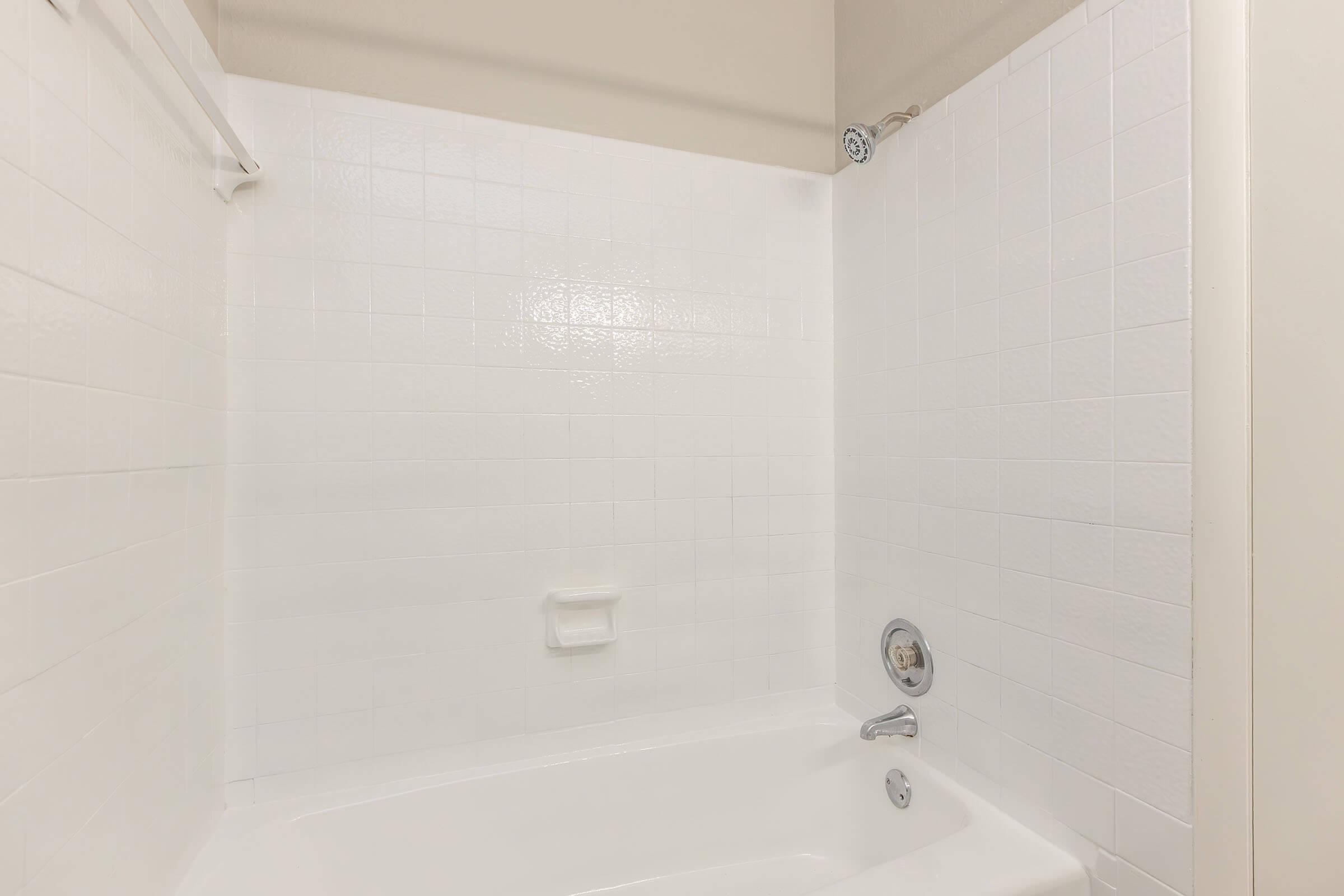 A clean, empty bathroom with a white tiled wall and a white bathtub. The showerhead is mounted on the wall, and there is a soap dish and a faucet visible. The overall appearance is bright and minimalist, creating a fresh and airy atmosphere.