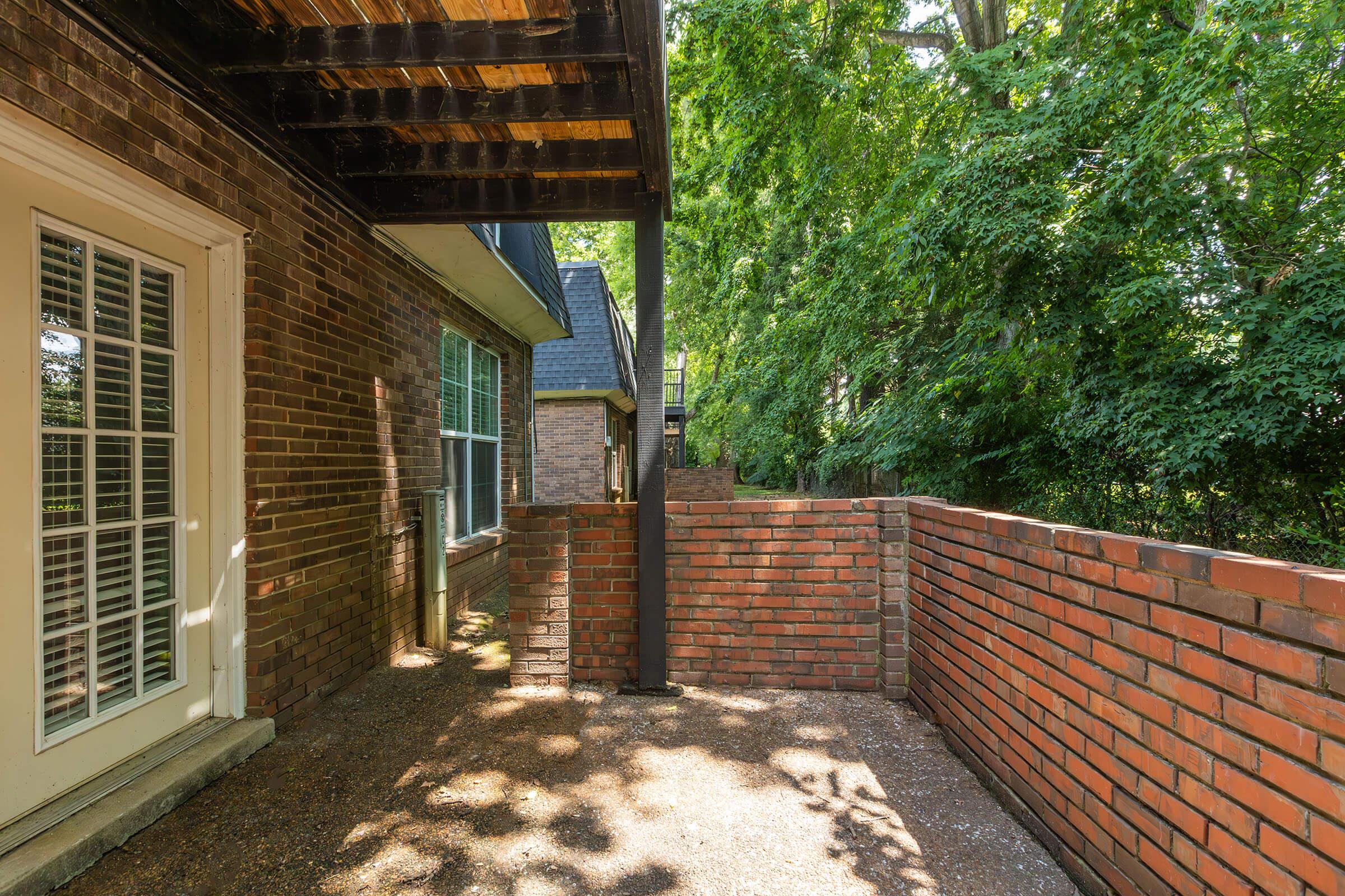 A corner view of a brick patio surrounded by greenery, with a wooden overhang above. A door with French-style windows leads into the house. The brick walls are partially covered by trees, creating a secluded outdoor space. Sunlight filters through the leaves, illuminating the area.