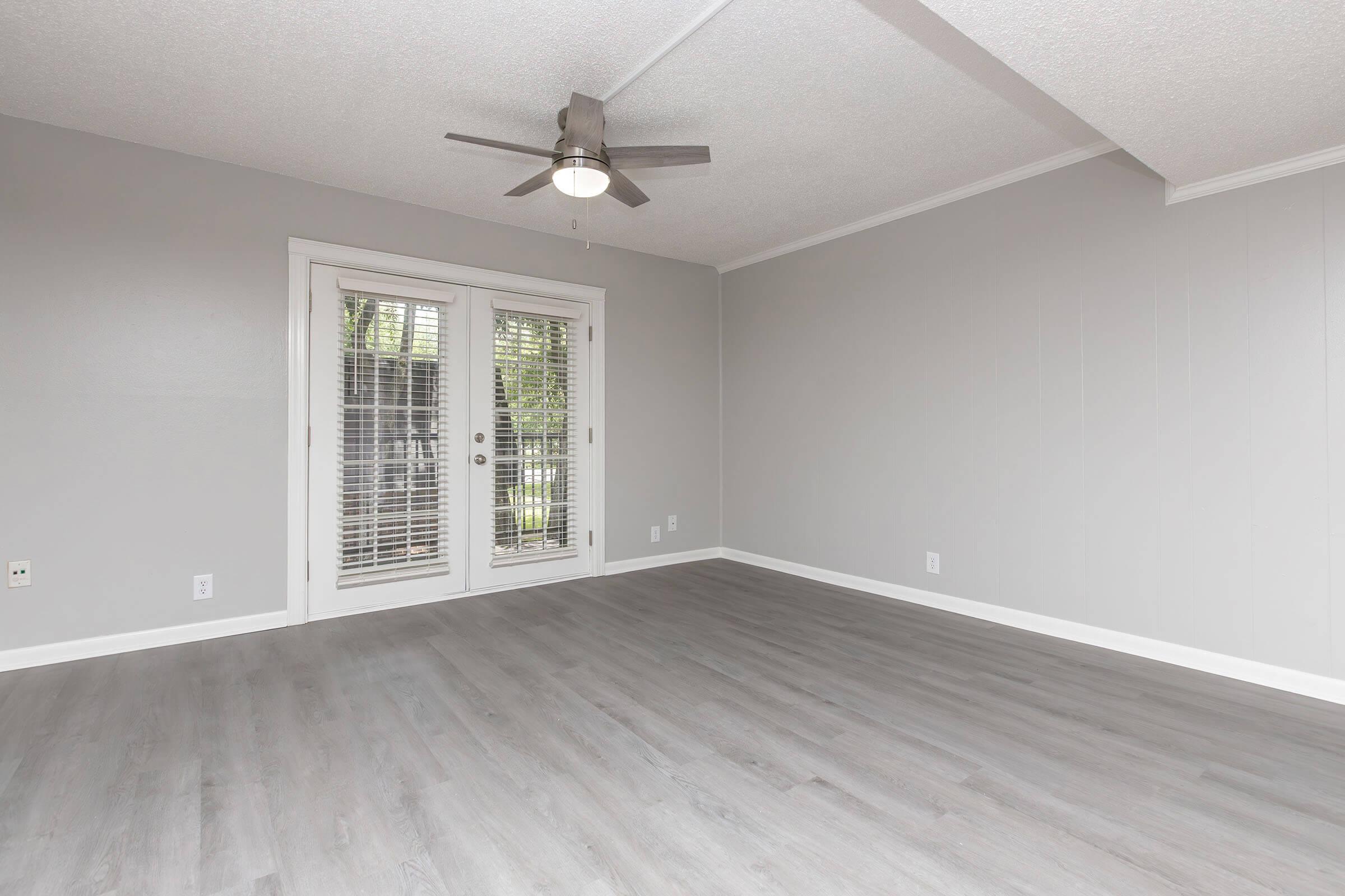 A spacious, empty living room with light gray walls and smooth, light-colored flooring. It features a ceiling fan and sliding glass doors with shutters that lead to an outdoor area. The room is well-lit, creating an inviting atmosphere.
