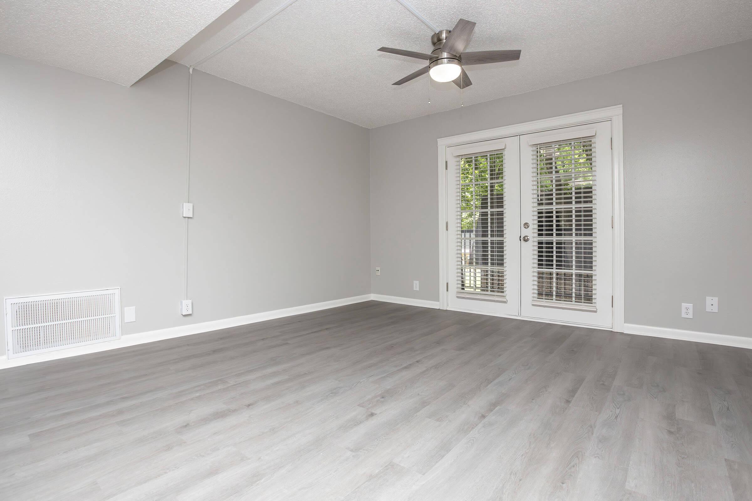 A spacious, empty room with light gray walls and a light wood-like floor. Features include a ceiling fan, a pair of French doors leading outside, and a wall vent. The room is well-lit, offering a clean slate for potential furniture arrangement.