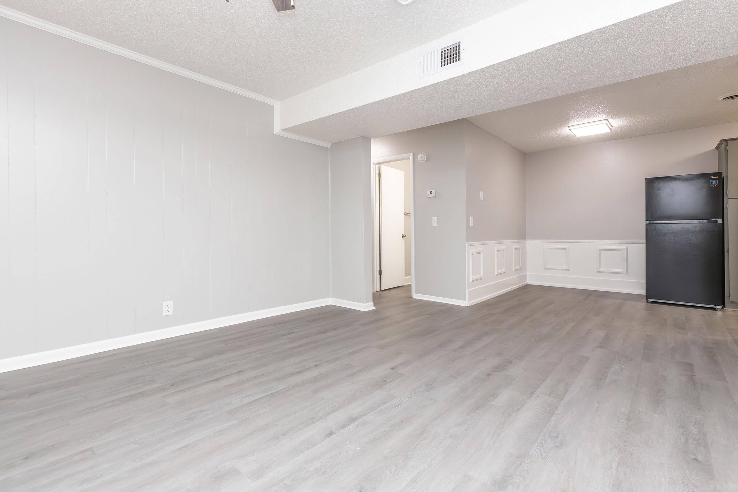 Interior of a modern, empty living space featuring light gray walls, new laminate flooring, and a black refrigerator. A doorway leads to another room, with a ceiling light fixture visible. The space has a clean, minimalist look with wainscoting along the lower part of the wall.