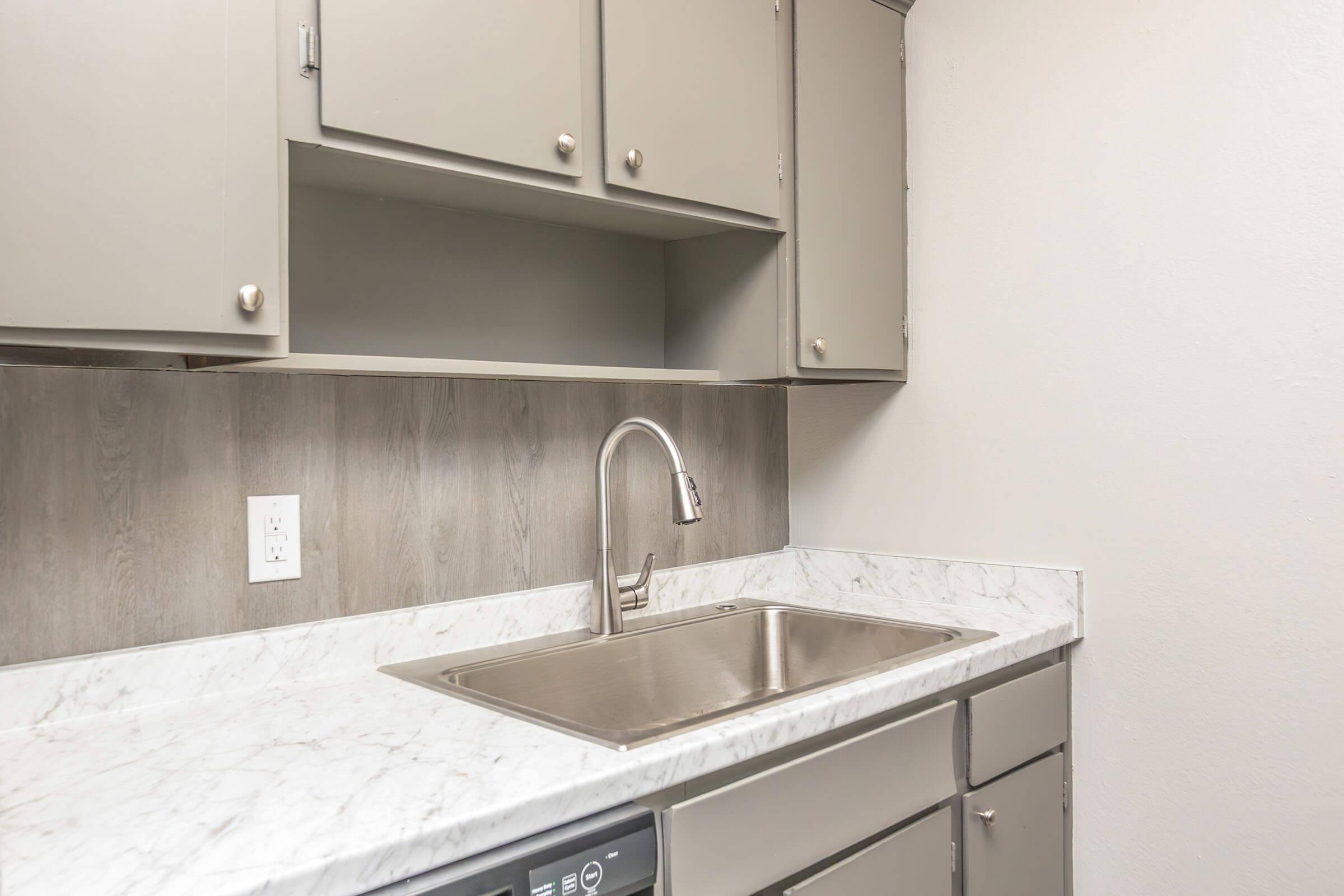 Modern kitchen area featuring a stainless steel sink, marble countertop, and light gray cabinets. The wall cabinet above the sink is empty, and there is a dark wood panel behind the countertop. The design is sleek and minimalist.