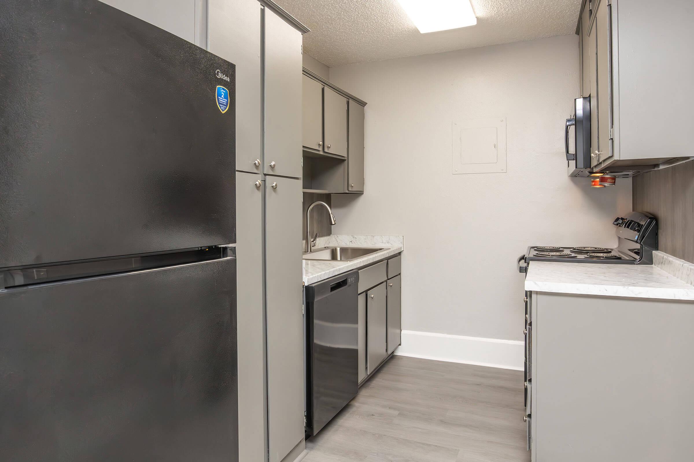 A modern kitchen featuring gray cabinets, a black refrigerator, a stainless steel sink, and a black stove. The countertops are light-colored, and the kitchen has laminate flooring. The walls are painted in a neutral tone, and there is overhead lighting for visibility.