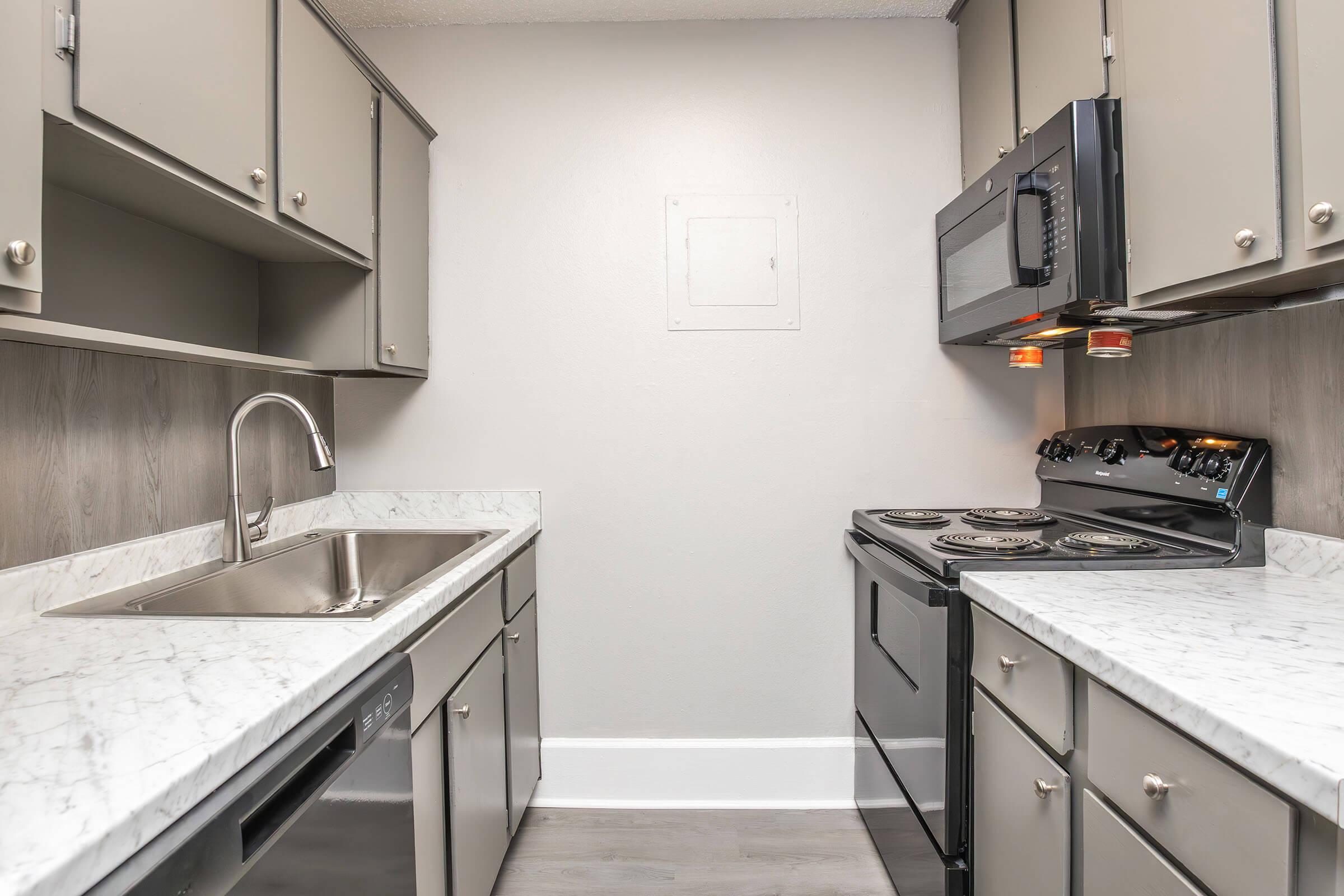 Modern kitchen with gray cabinetry, stainless steel sink, and marble countertops. Includes a black stove, microwave, and dishwasher. The walls are painted in a neutral tone, and there is a small white access panel on the wall. The floor features a wood-like finish.