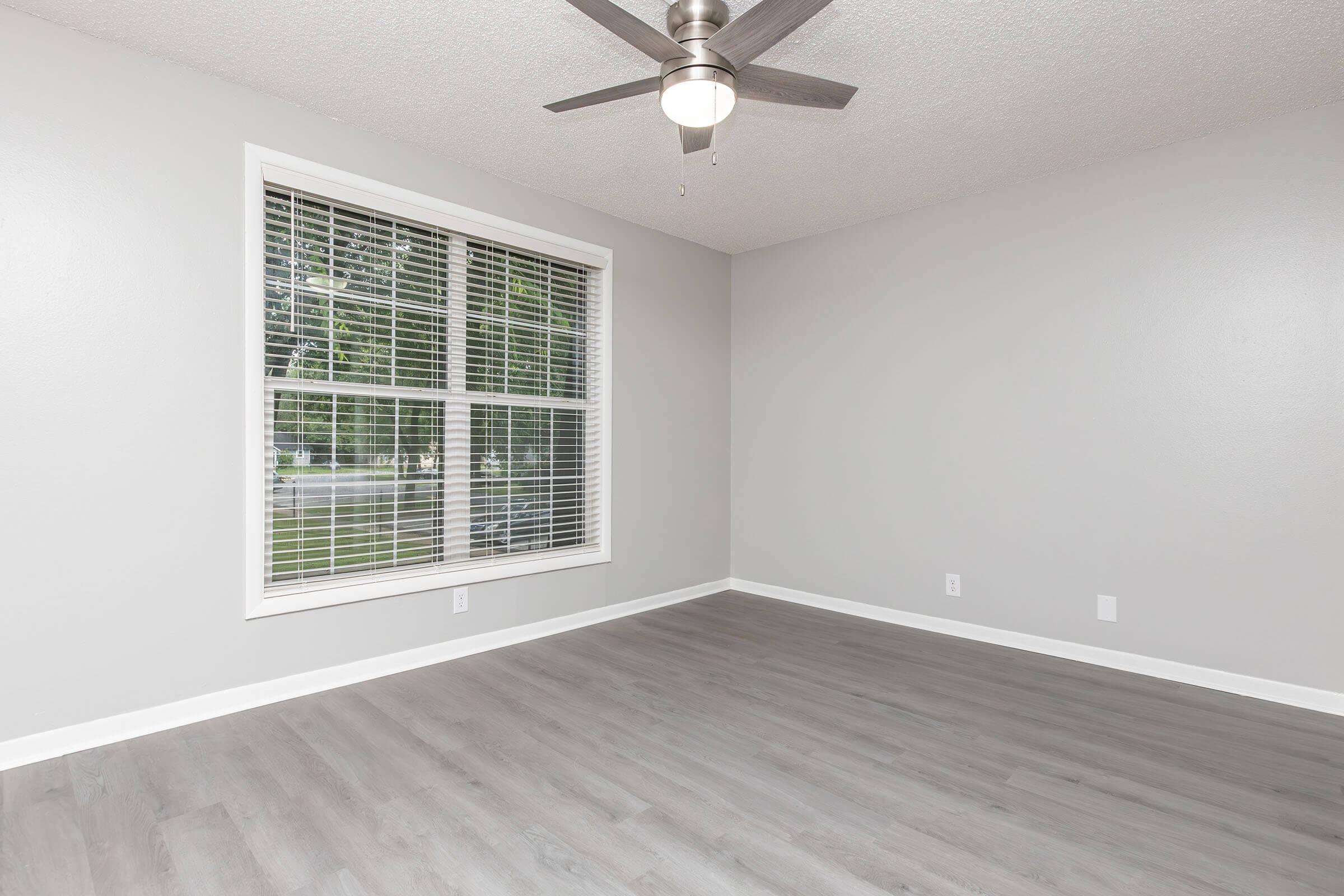 A bright, empty room with gray walls and light gray flooring. A large window with blinds allows natural light in, and a modern ceiling fan is mounted above. The space is uncluttered and has a clean, minimalist appearance, ideal for various decorative styles.