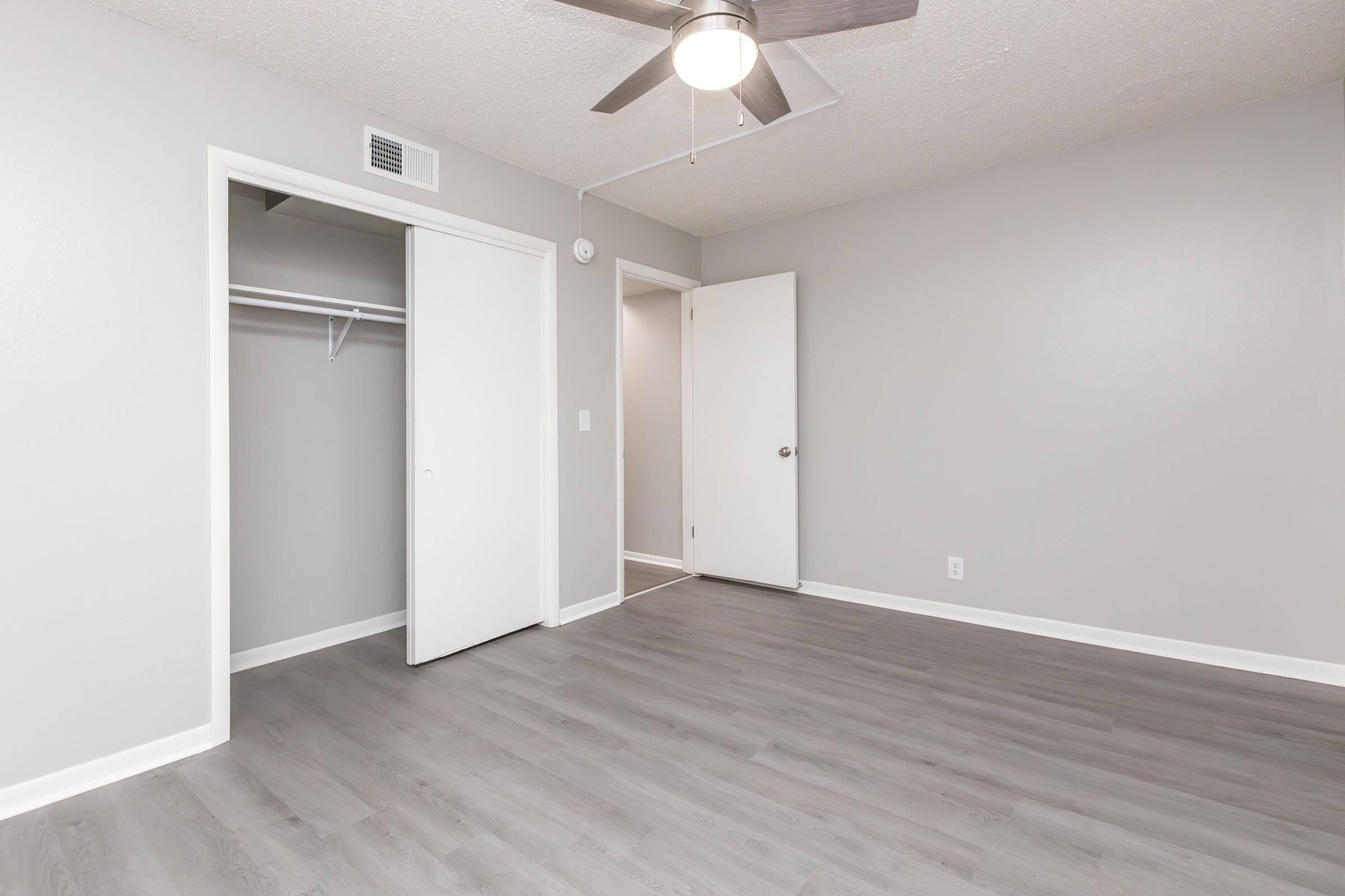 A spacious and well-lit room featuring gray walls and a light-colored wood floor. There is a ceiling fan and two doors: one leading to a closet and another to a hallway. The room appears freshly painted and is empty, providing a clean, modern aesthetic.