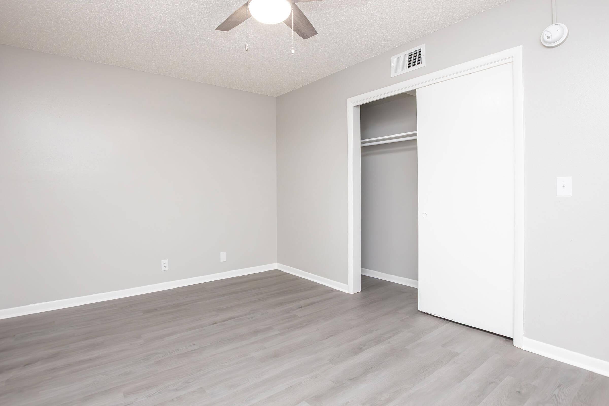 A minimally decorated room featuring light gray walls and wooden flooring. A ceiling fan hangs from the ceiling, and there's a white closet with sliding doors. The space appears bright and uncluttered, suitable for various uses such as a bedroom or office.