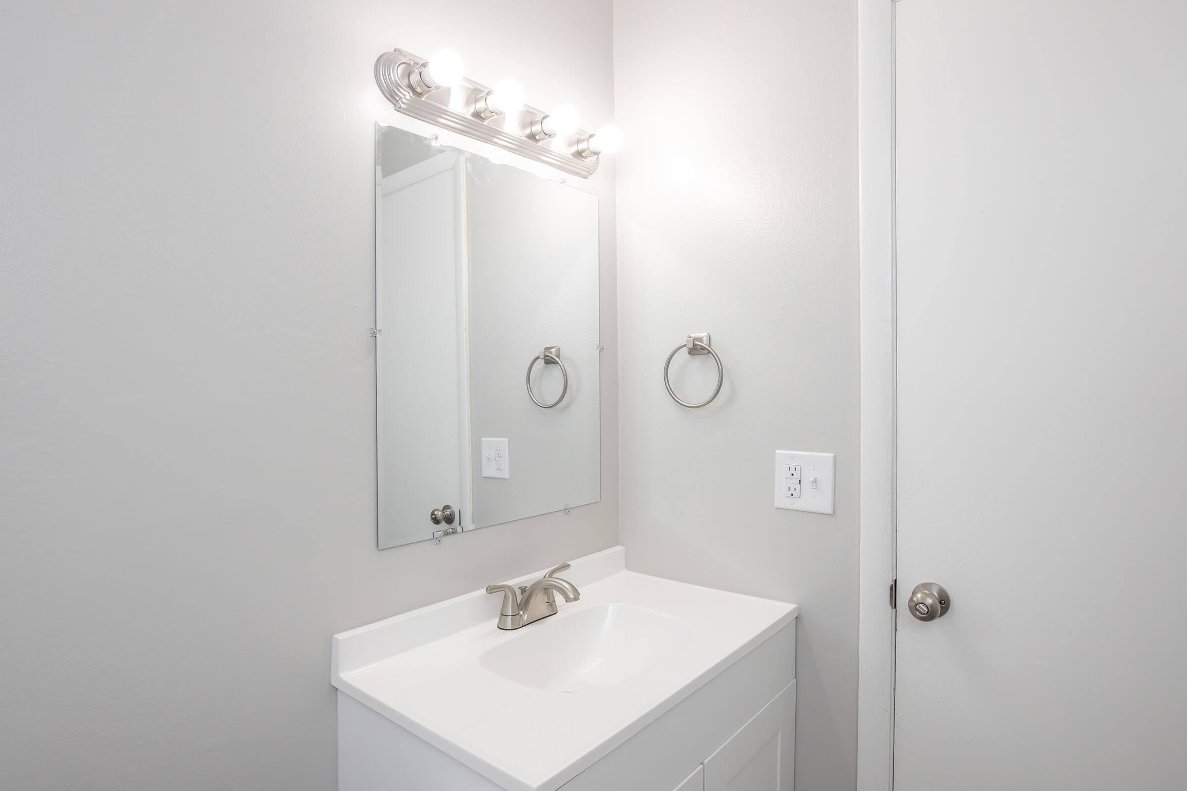 A modern bathroom featuring a white vanity with a sink, a large mirror above it, and a light fixture with several bulbs. A towel ring is mounted on the wall beside the mirror, and a door is visible on the right side. The walls are painted in a light gray color, contributing to a clean and bright appearance.