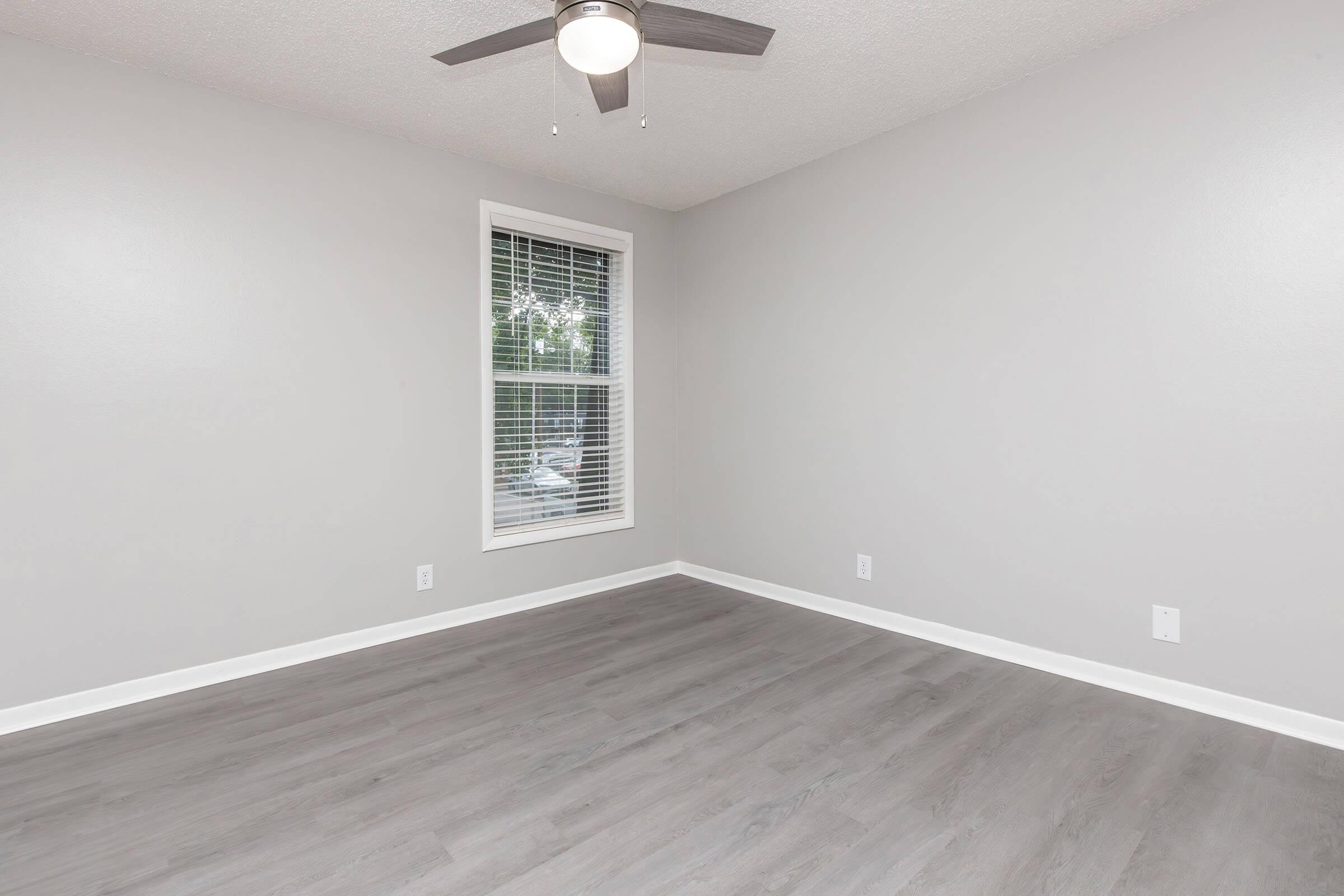 A well-lit, empty room with light gray walls and a ceiling fan. The floor is covered in light-colored laminate, and there is a window with blinds allowing natural light to enter. The space has a clean and modern appearance, ideal for various uses.