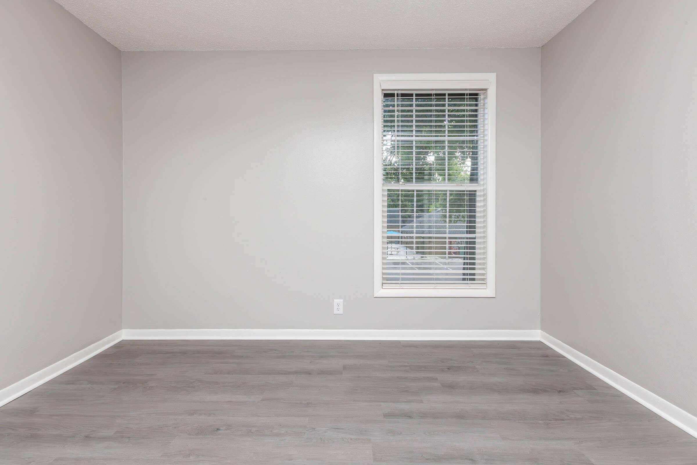 Empty room with light gray walls and a window featuring white blinds. The floor is light-colored laminate. The space is minimalistic, devoid of furniture, creating a bright and airy atmosphere. Natural light enters through the window, illuminating the room.