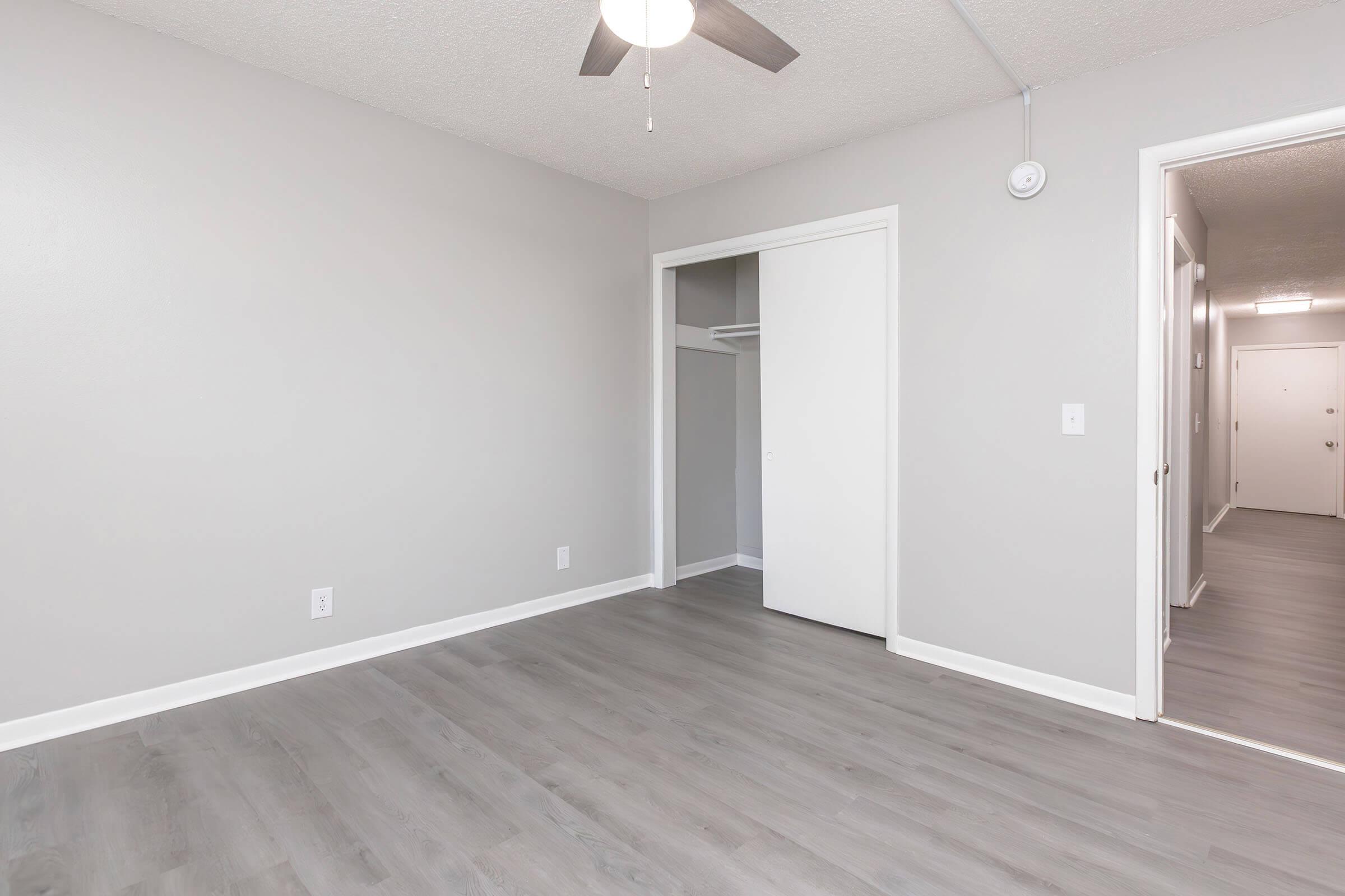 A room with light gray walls and a ceiling fan. The floor is covered in light wood-like laminate. There is an open closet with sliding doors, and a hallway is visible in the background leading to other rooms. The space is empty, offering a blank canvas for decoration.