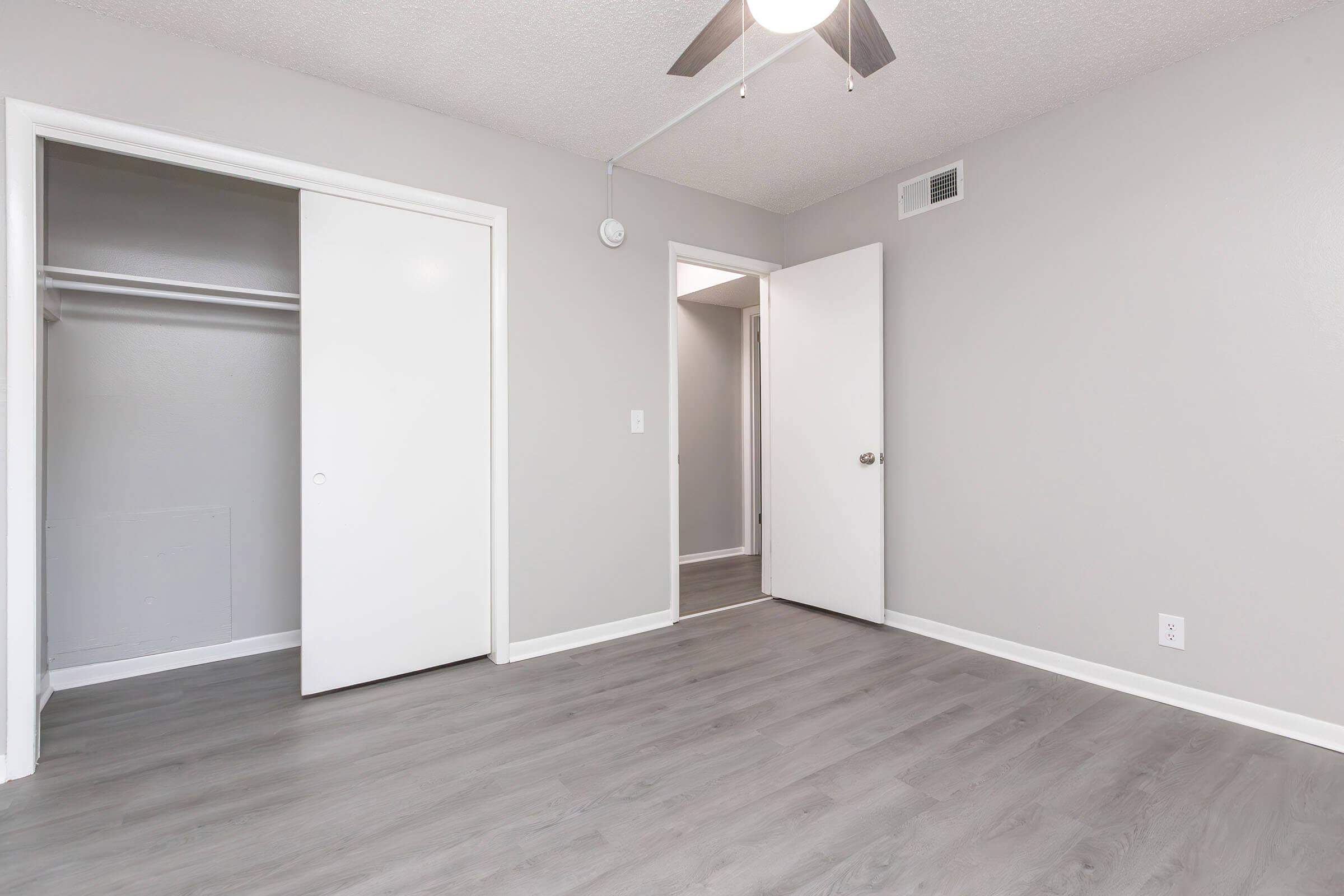 A spacious, well-lit room featuring light gray walls, a ceiling fan, and laminate flooring. There are two doors: one leading to a small closet and another to an adjoining space. The room is unadorned, creating a blank canvas for personalization.