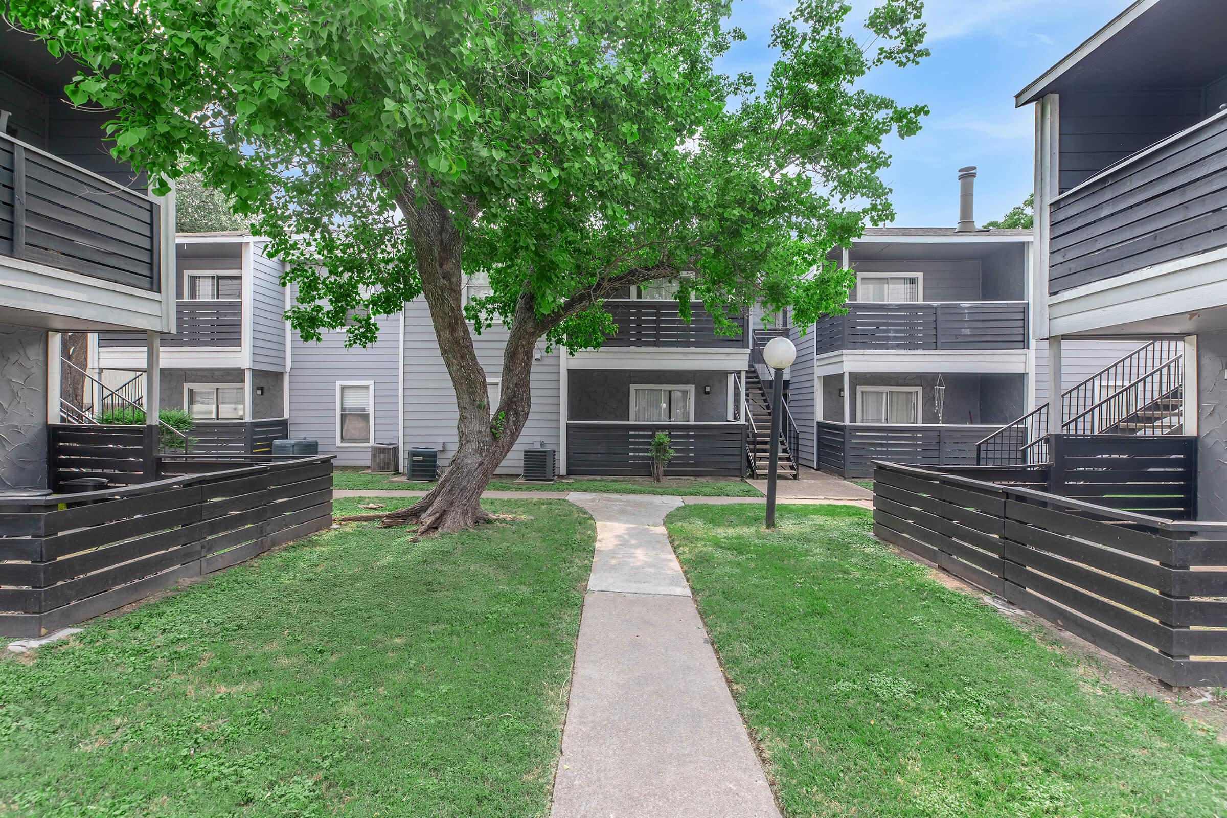 A landscaped courtyard surrounded by two-story apartment buildings. Grass covers the ground, and a tree provides shade. The buildings have black balconies and a central pathway leading through the space, with light fixtures and greenery enhancing the outdoor environment.