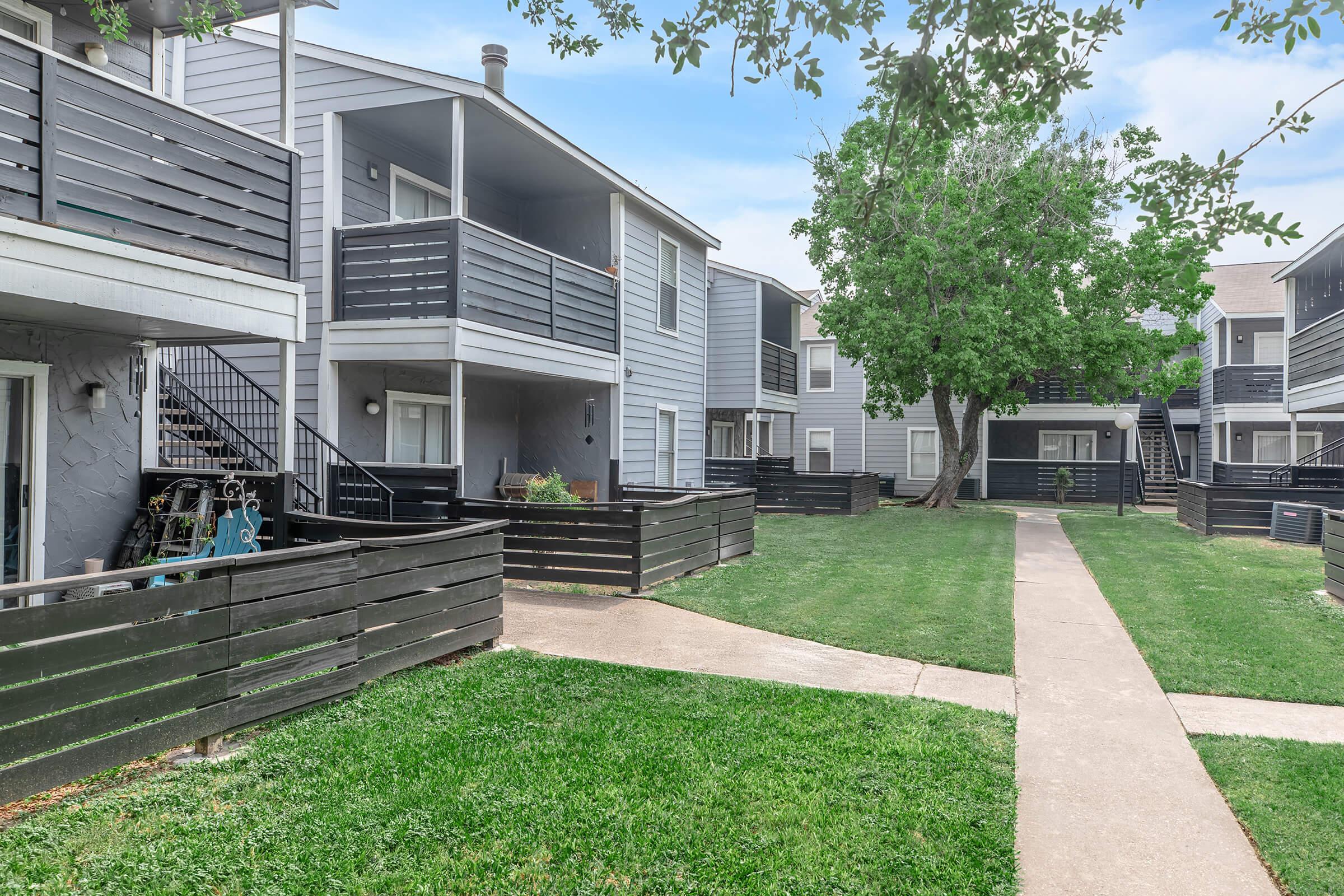 A view of a residential complex featuring two-story apartment buildings with gray siding, black railings, and balconies. The landscape includes a well-maintained grassy area and a concrete pathway leading through the complex. Trees provide shade, enhancing the inviting atmosphere of the community.