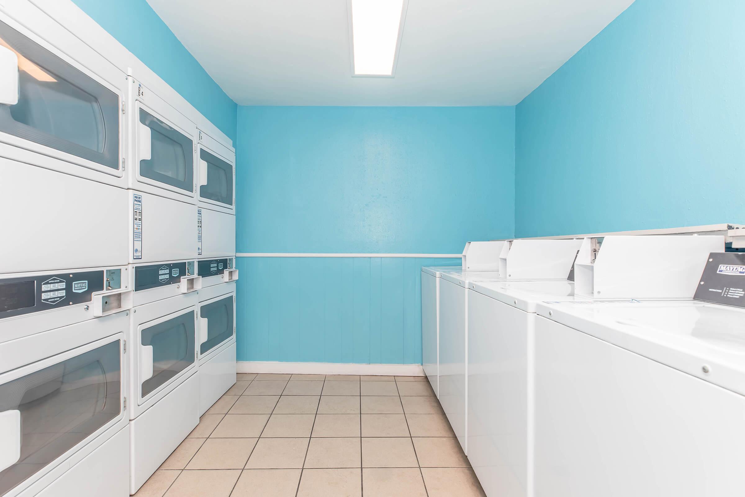 A clean and well-lit laundry room featuring multiple stacked dryers and washers. The walls are painted a vibrant blue, and the tiled floor is neutral. The space is organized, with machines positioned neatly along the walls, creating an efficient laundry area.