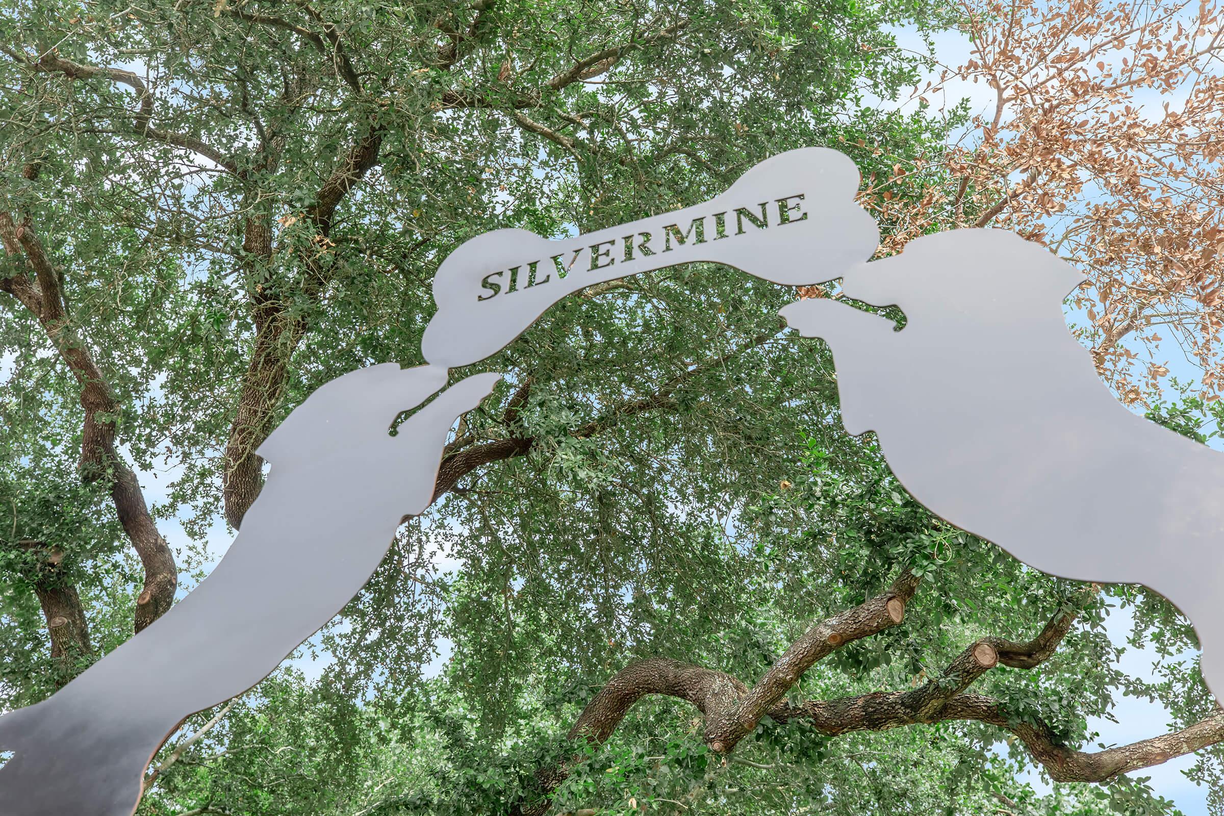 A metal sign featuring the word "SILVERMINE" suspended between two silhouettes of dogs, surrounded by lush trees and greenery. The sign is positioned above, with branches in the background, creating a welcoming entrance to a pet-related area or park.